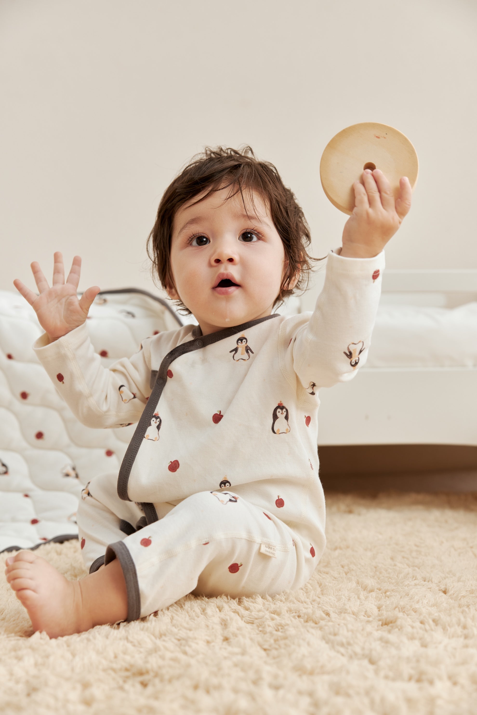 Baby in a white kimono sleeper with penguin design holding a wooden toy on a carpeted floor.