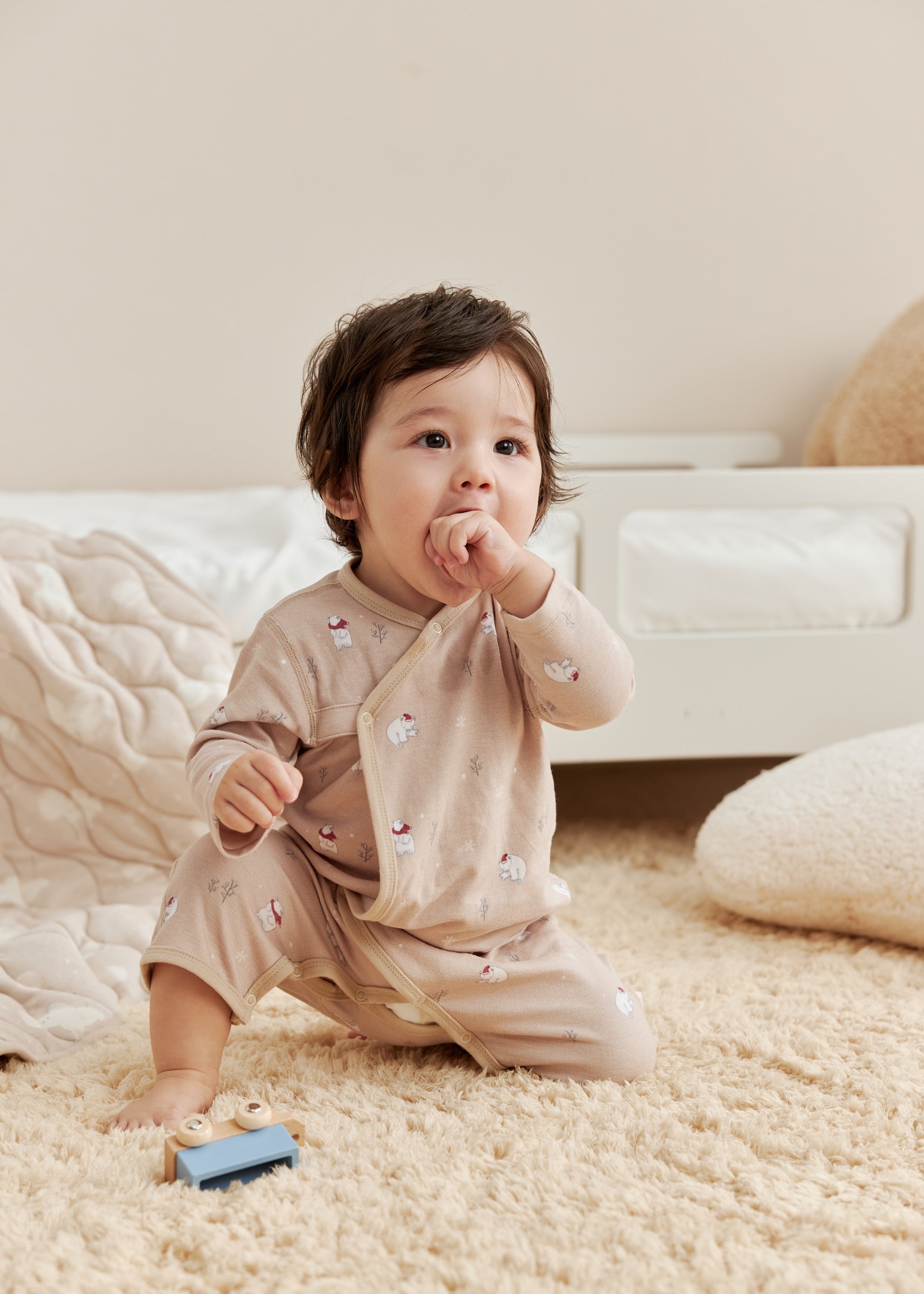 Baby in a beige outfit standing on a carpeted floor with a neutral background