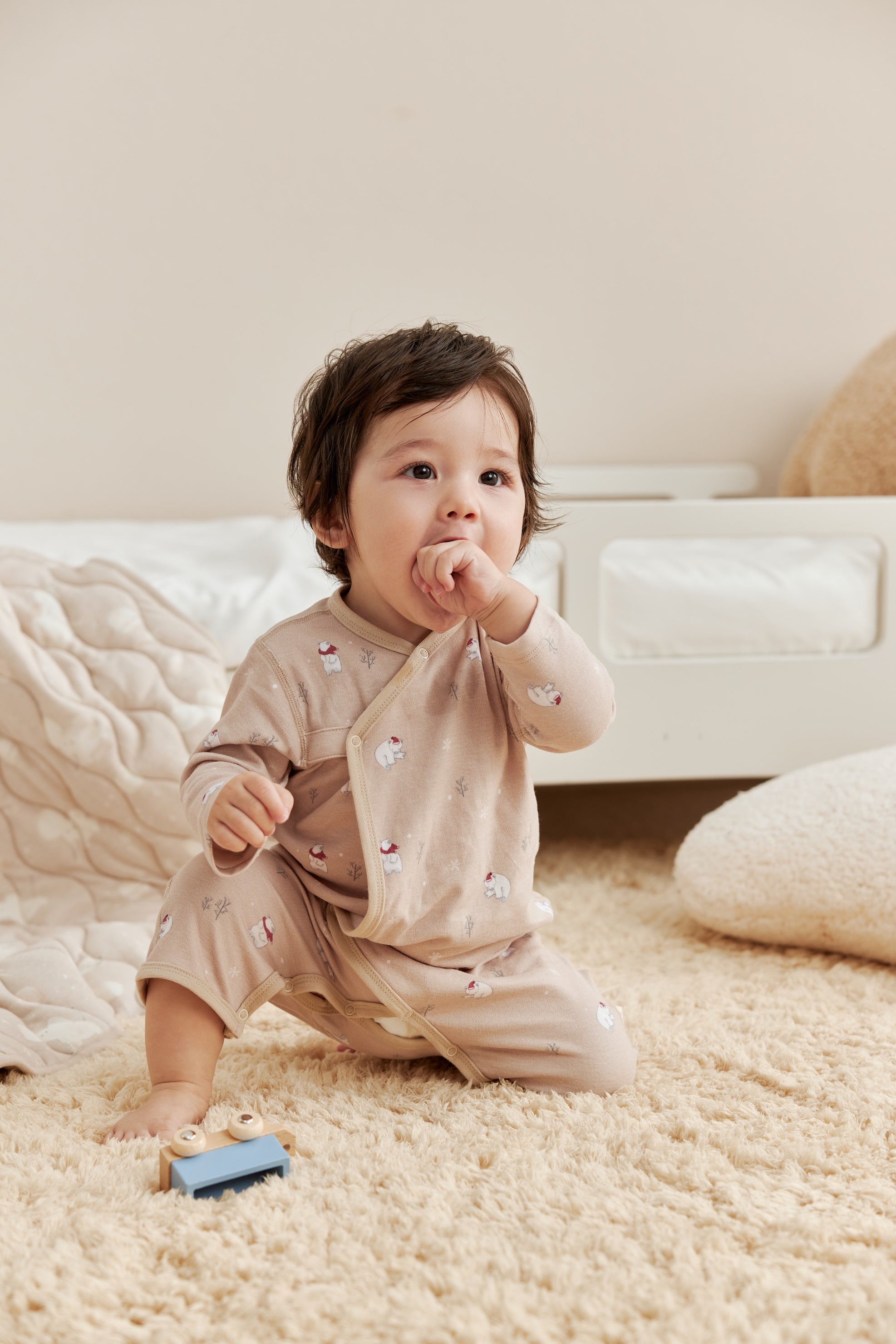 Baby in a beige outfit standing on a carpeted floor with a neutral background