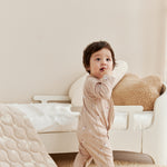 Baby in a beige onesie standing in a room with a white chair and textured rug.