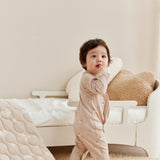 Baby in a beige onesie standing in a room with a white chair and textured rug.