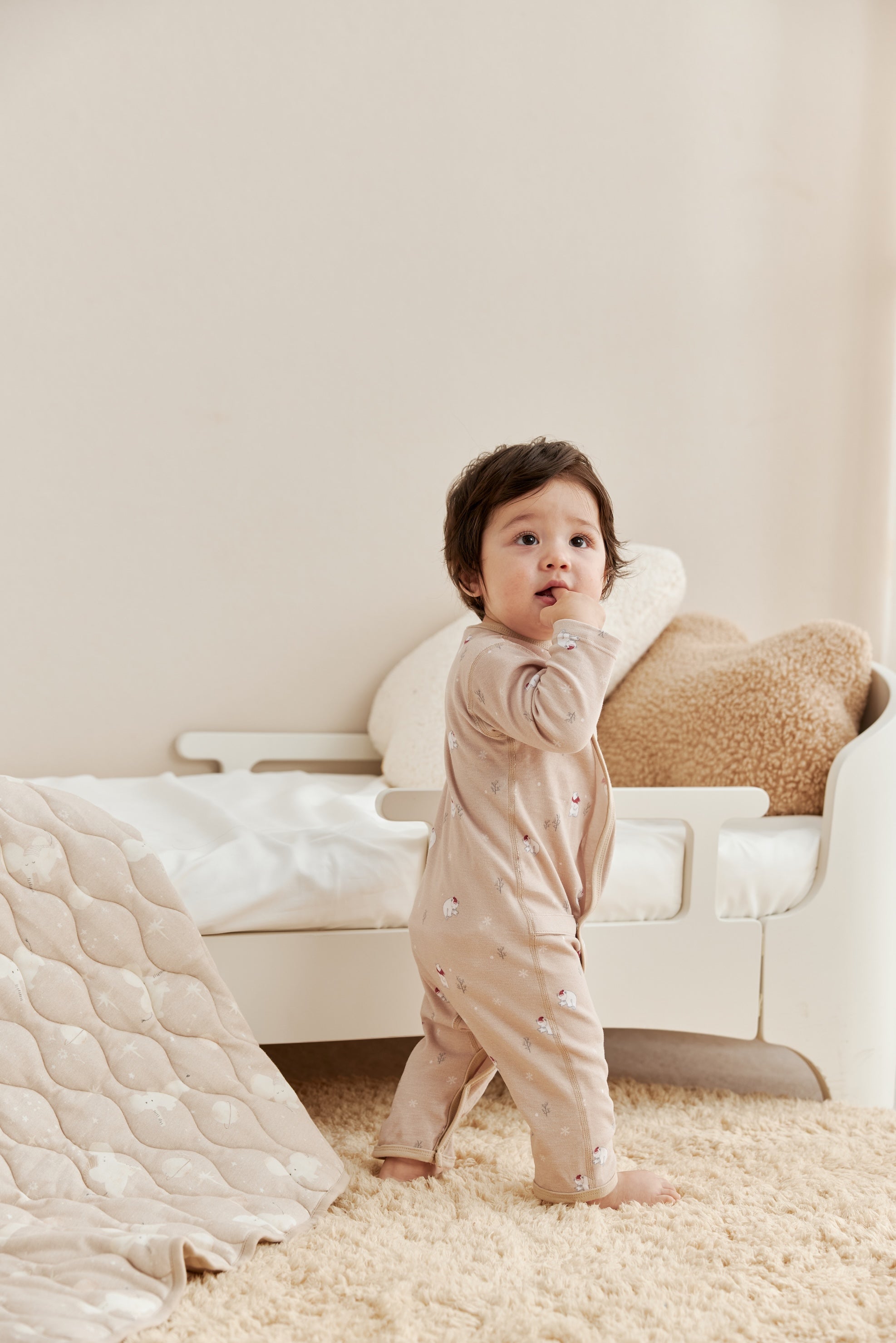 Baby in a beige onesie standing in a room with a white chair and textured rug.