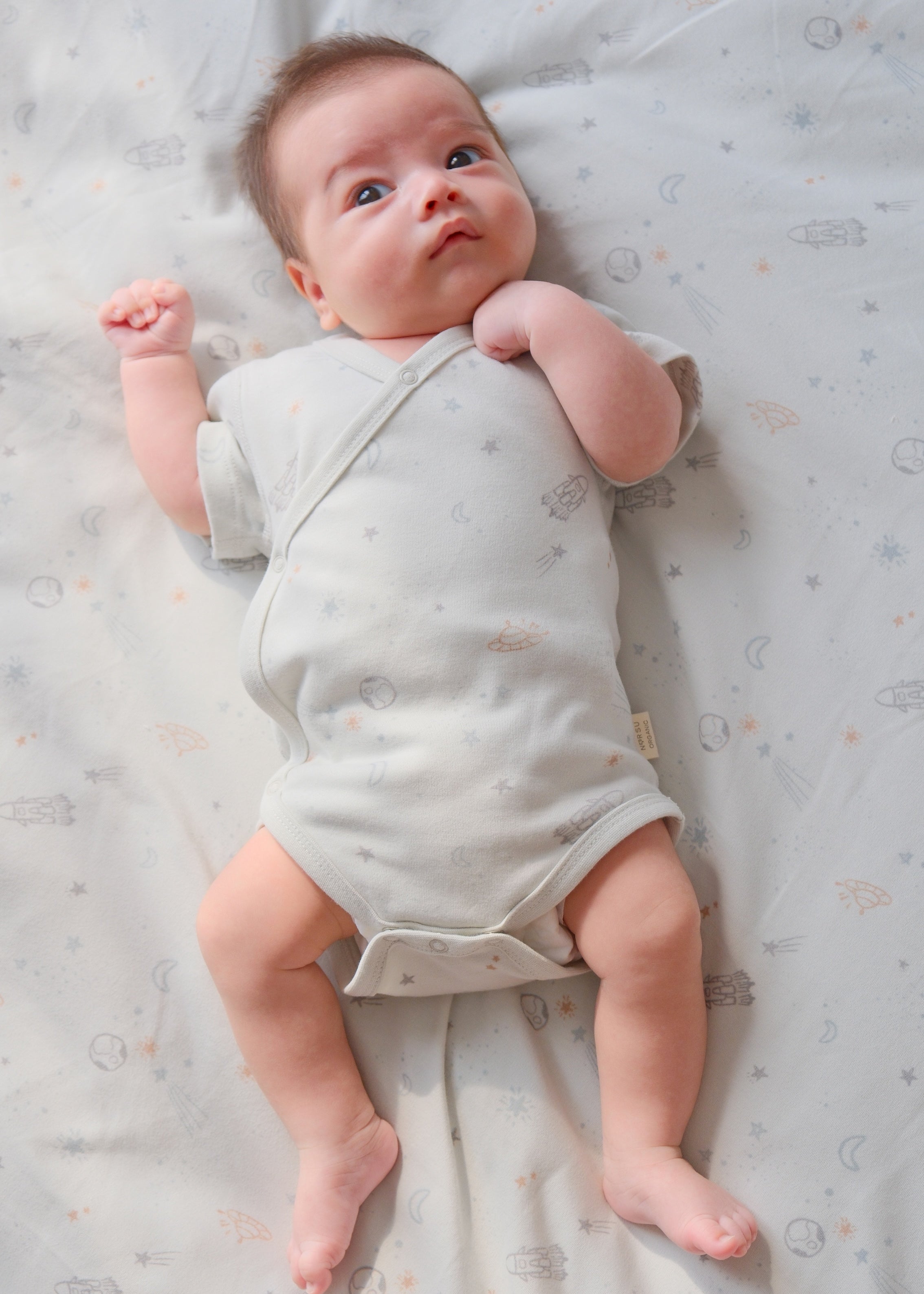 1. A peaceful baby  on a bed, dressed in a cute organic kimono onesie with a space theme.
