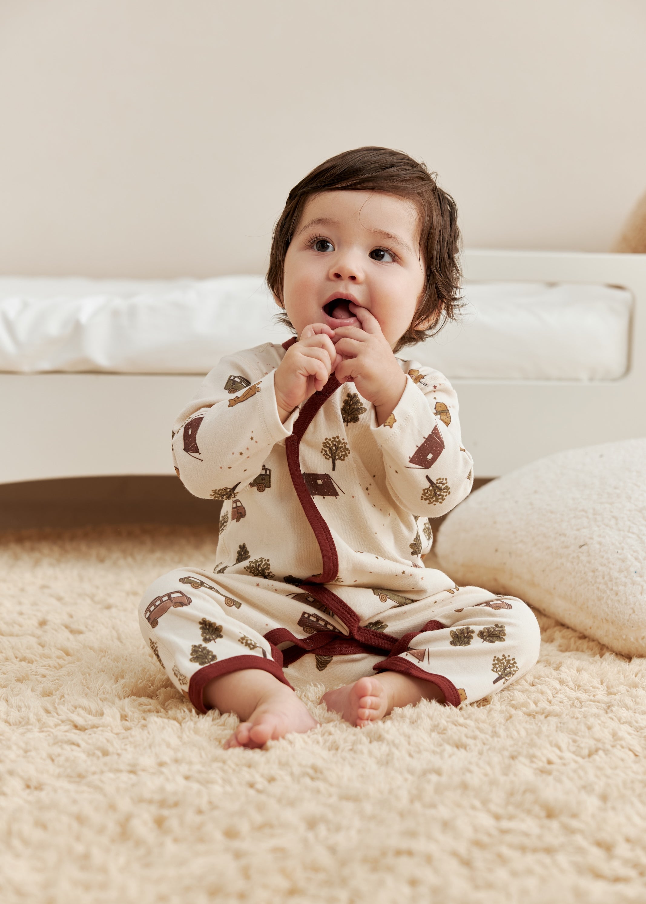 Baby in a patterned sleeper sitting on a carpeted floor.