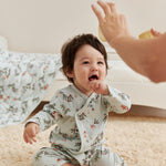 Baby in jolly sleeper sitting on a carpeted floor, reaching out towards an adult's hand.