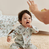 Baby in jolly sleeper sitting on a carpeted floor, reaching out towards an adult's hand.