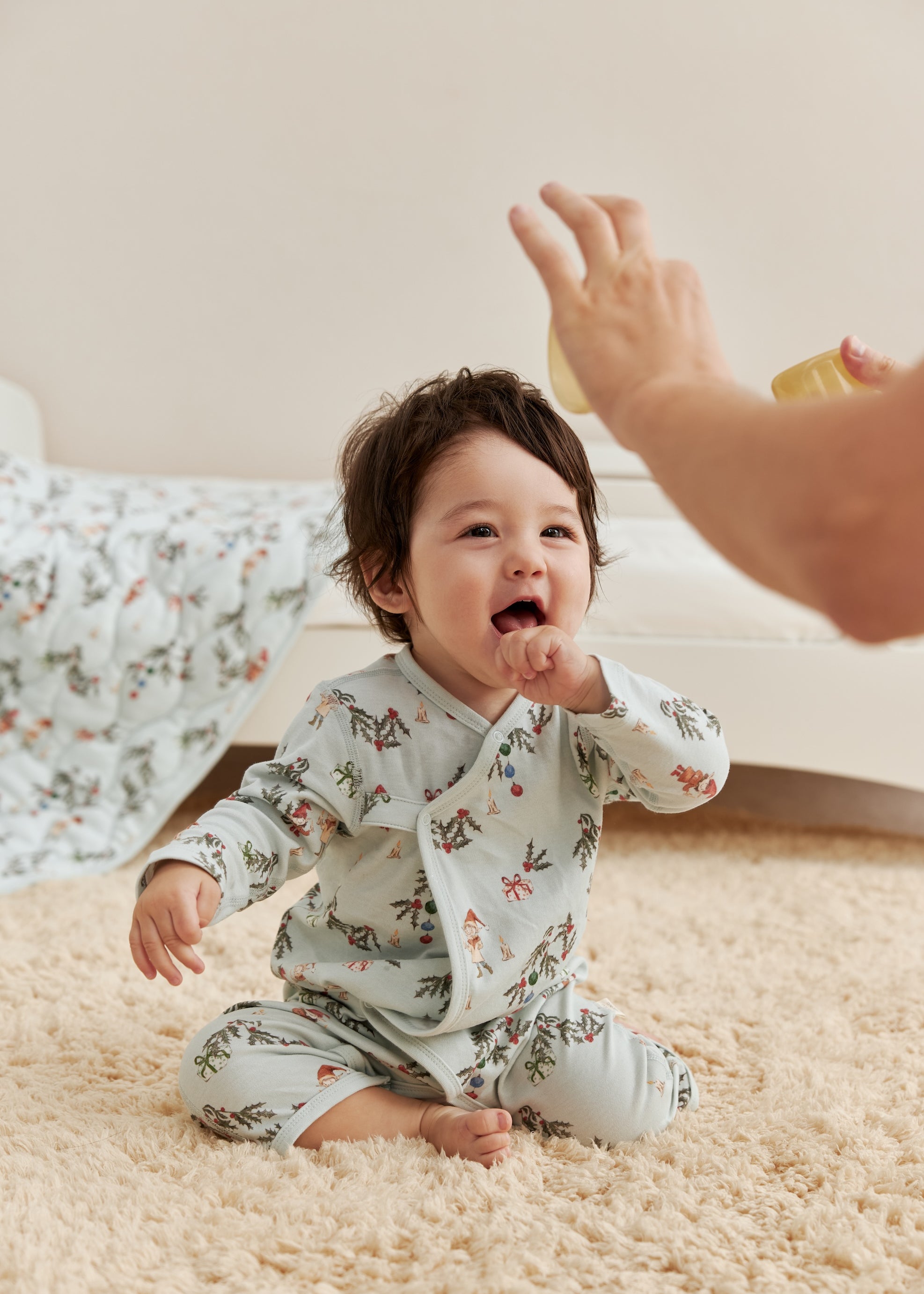 Baby in jolly sleeper sitting on a carpeted floor, reaching out towards an adult's hand.