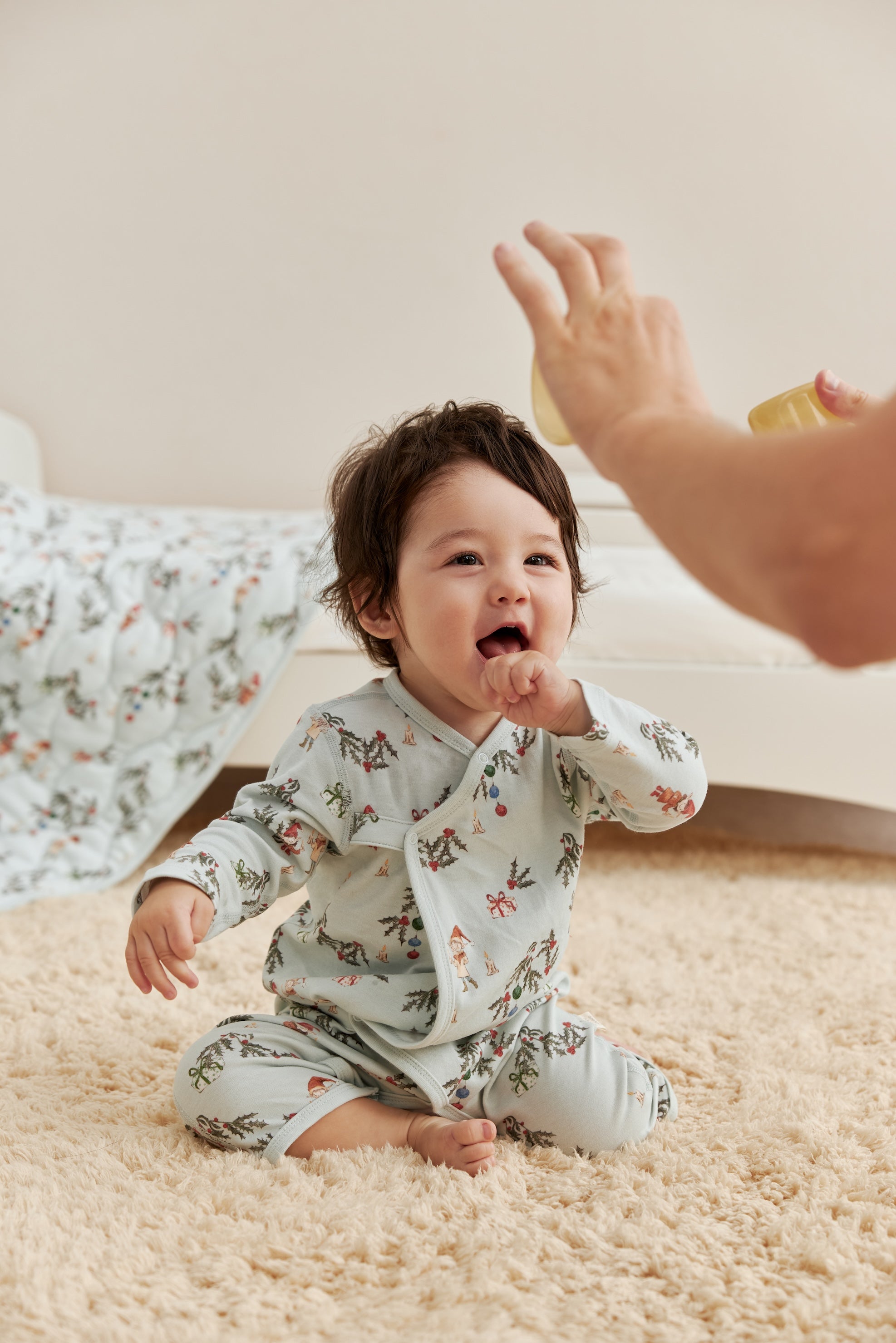 Baby in jolly sleeper sitting on a carpeted floor, reaching out towards an adult's hand.