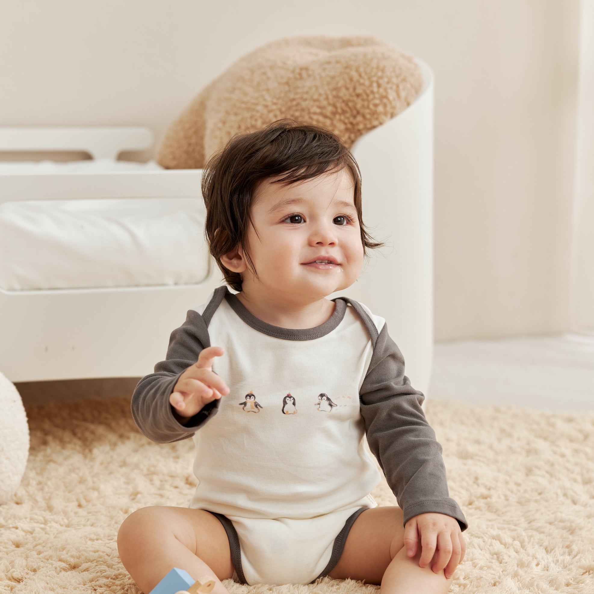 Baby sitting on a carpeted floor with a toy, wearing a gray and white onesie.