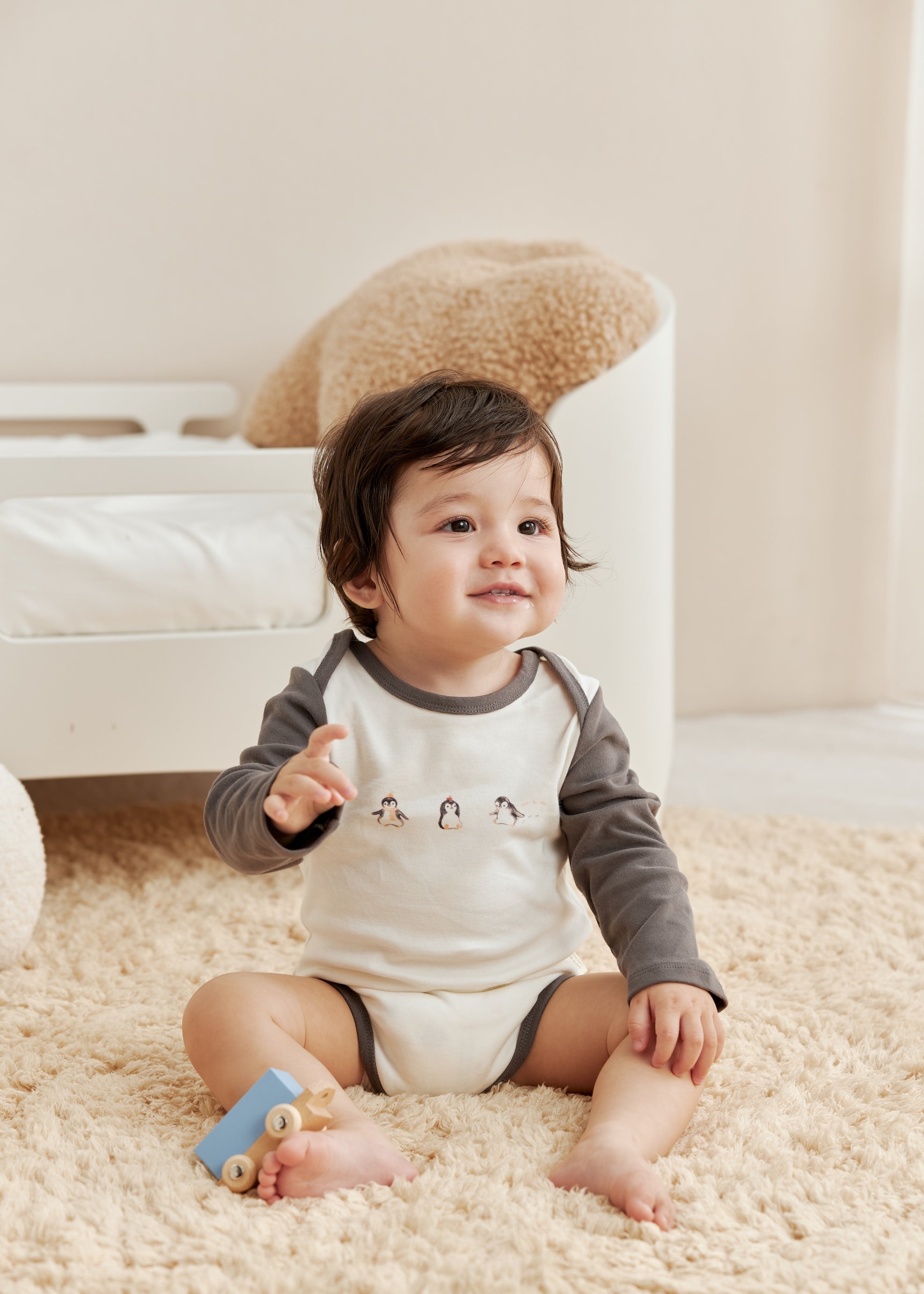 Baby sitting on a carpeted floor with a toy, wearing a gray and white onesie.