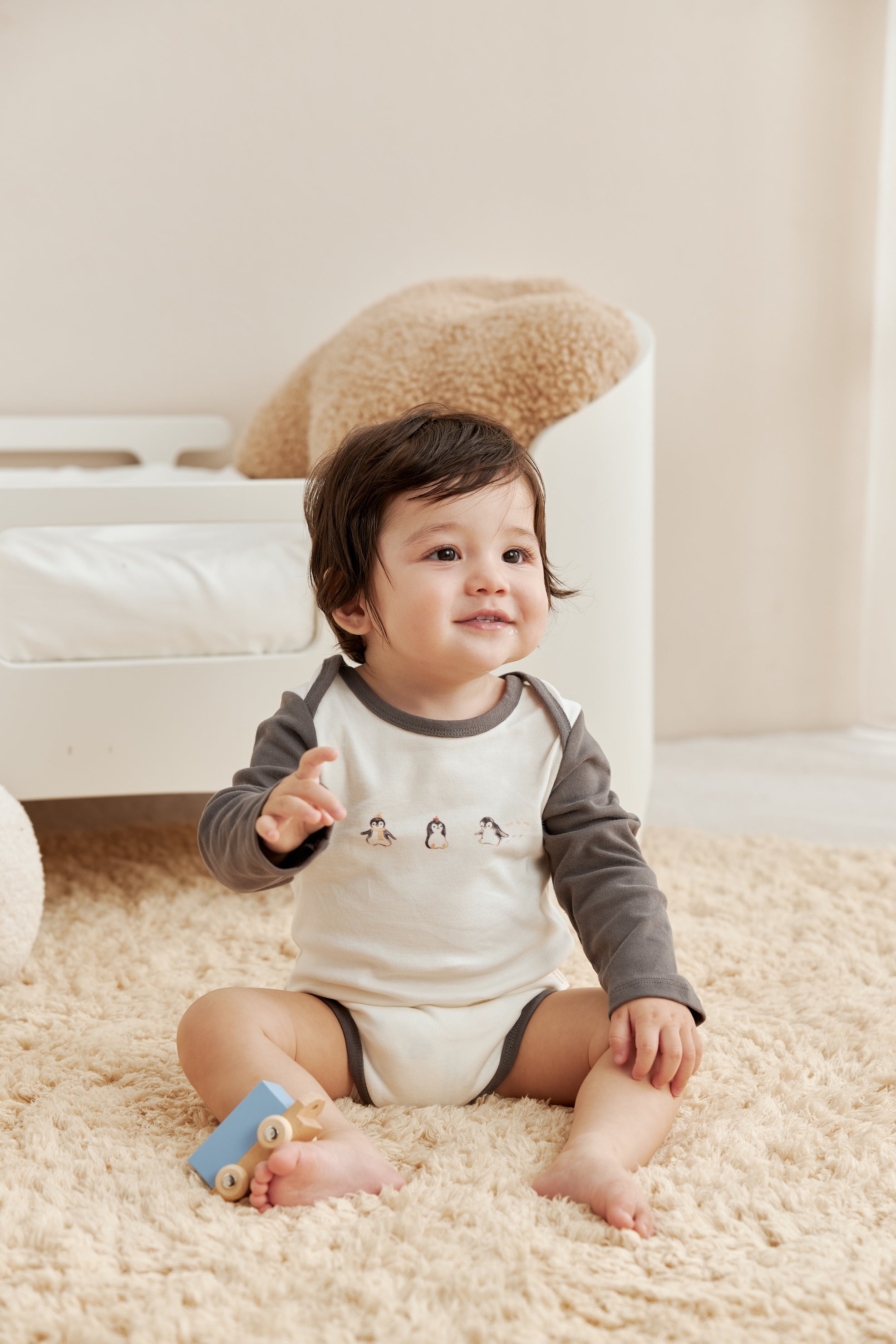 Baby sitting on a carpeted floor with a toy, wearing a gray and white onesie.
