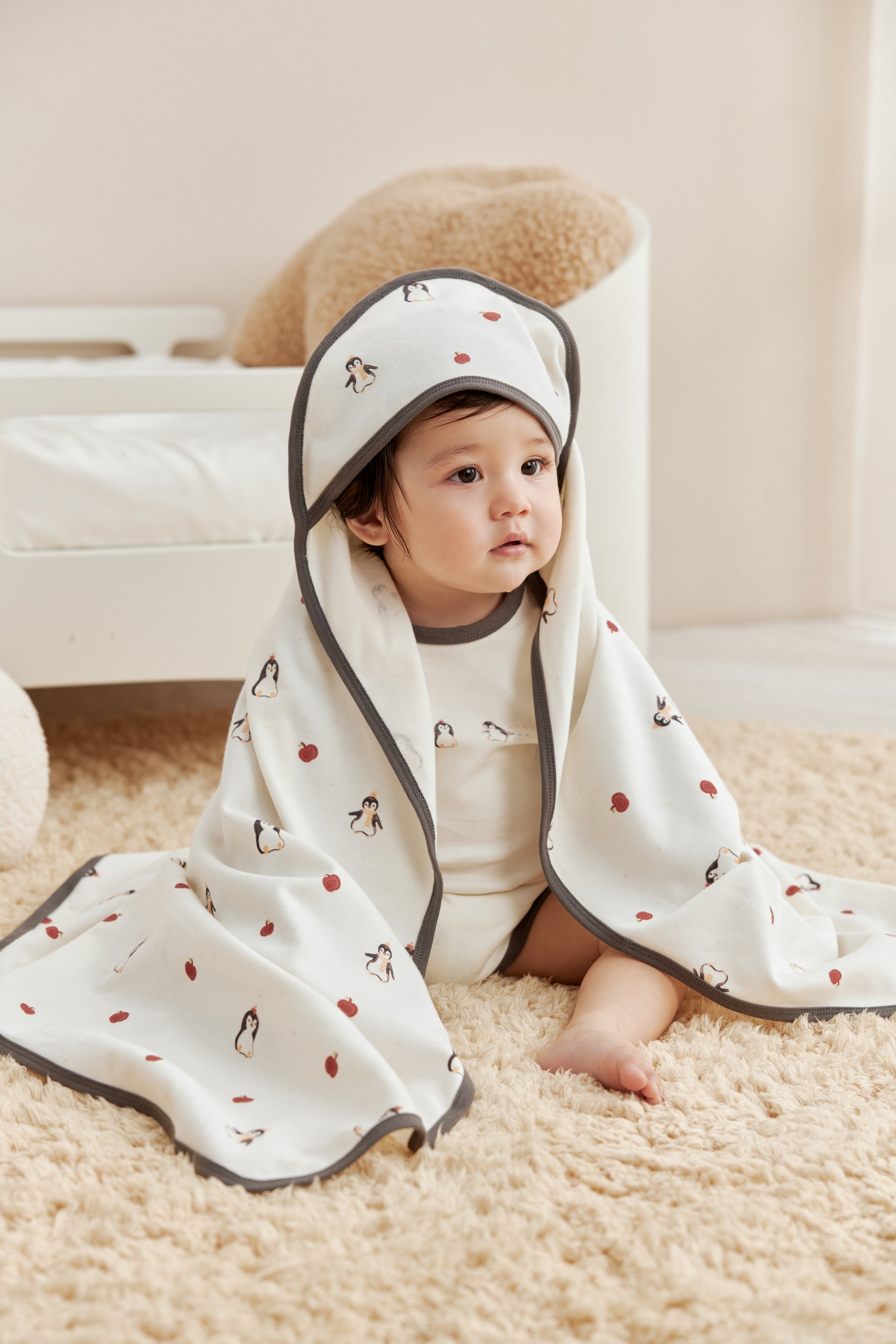 Baby wrapped in a white blanket with penguin patterns on a beige carpet.