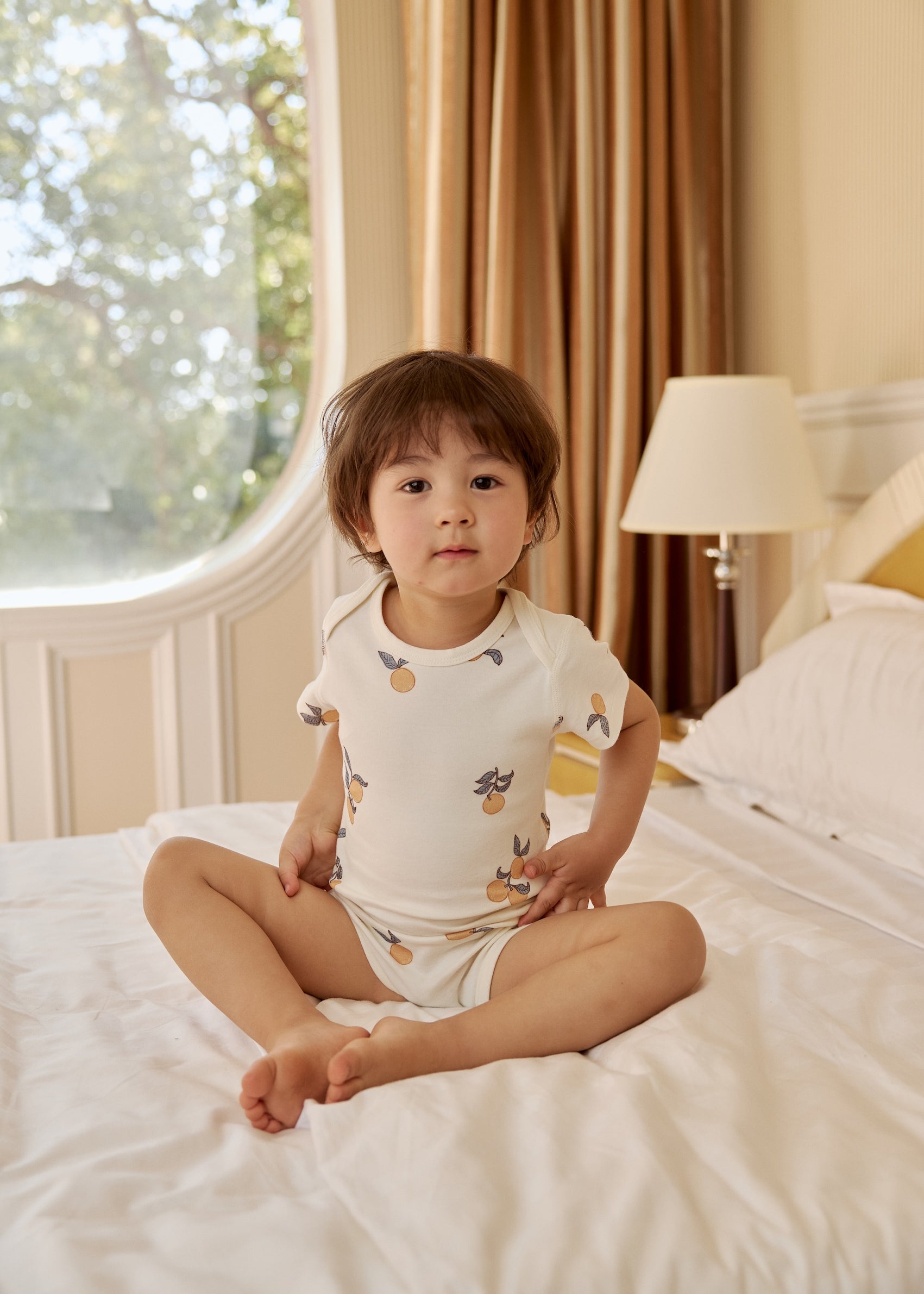 1. A young child sits on a bed, wearing a cute Baby Organic Short-Sleeve Onesie in a vibrant clementine color.
