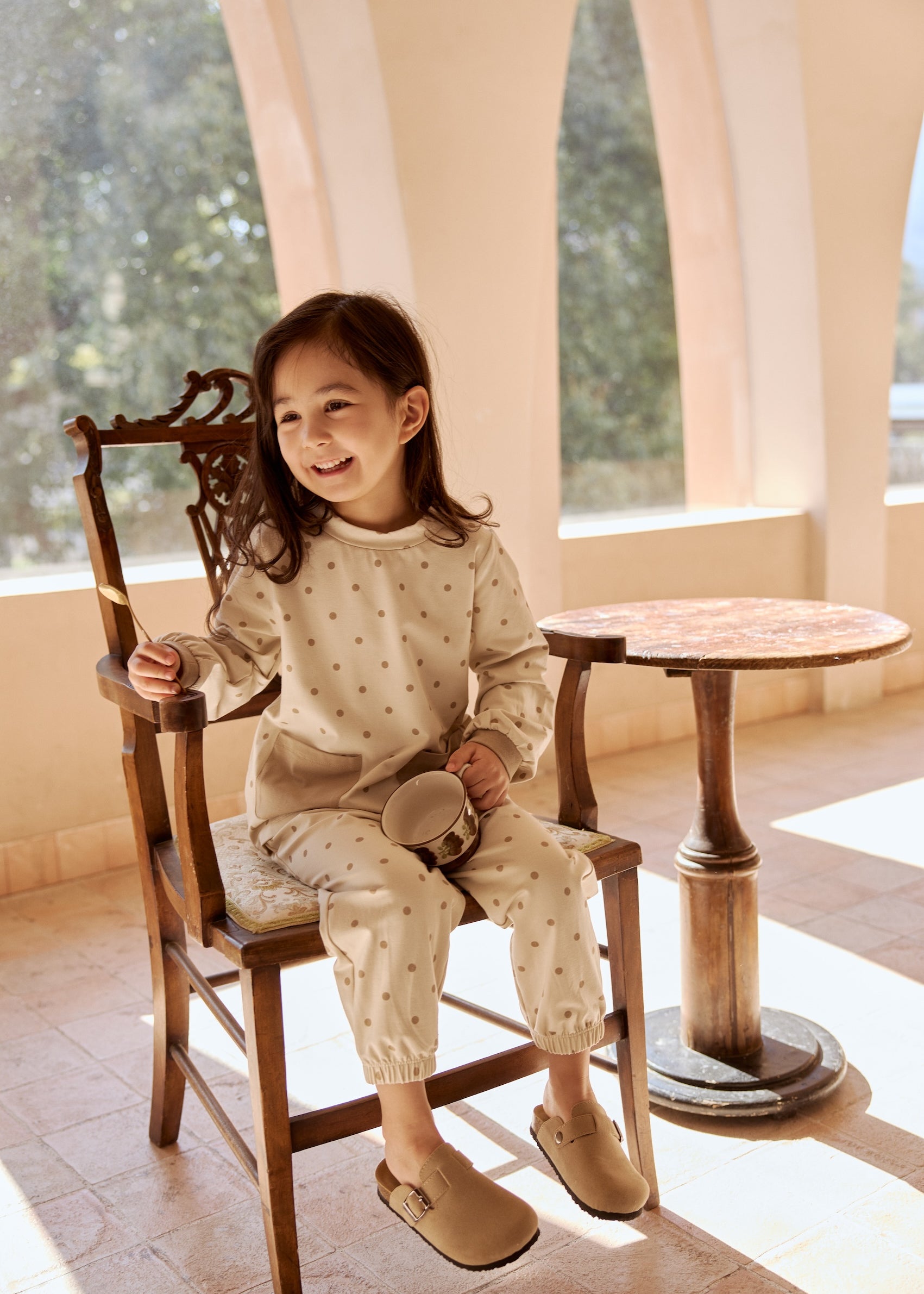 A little girl in a dot-patterned bamboo shirt sits on a chair, smiling in a cozy room filled with soft light