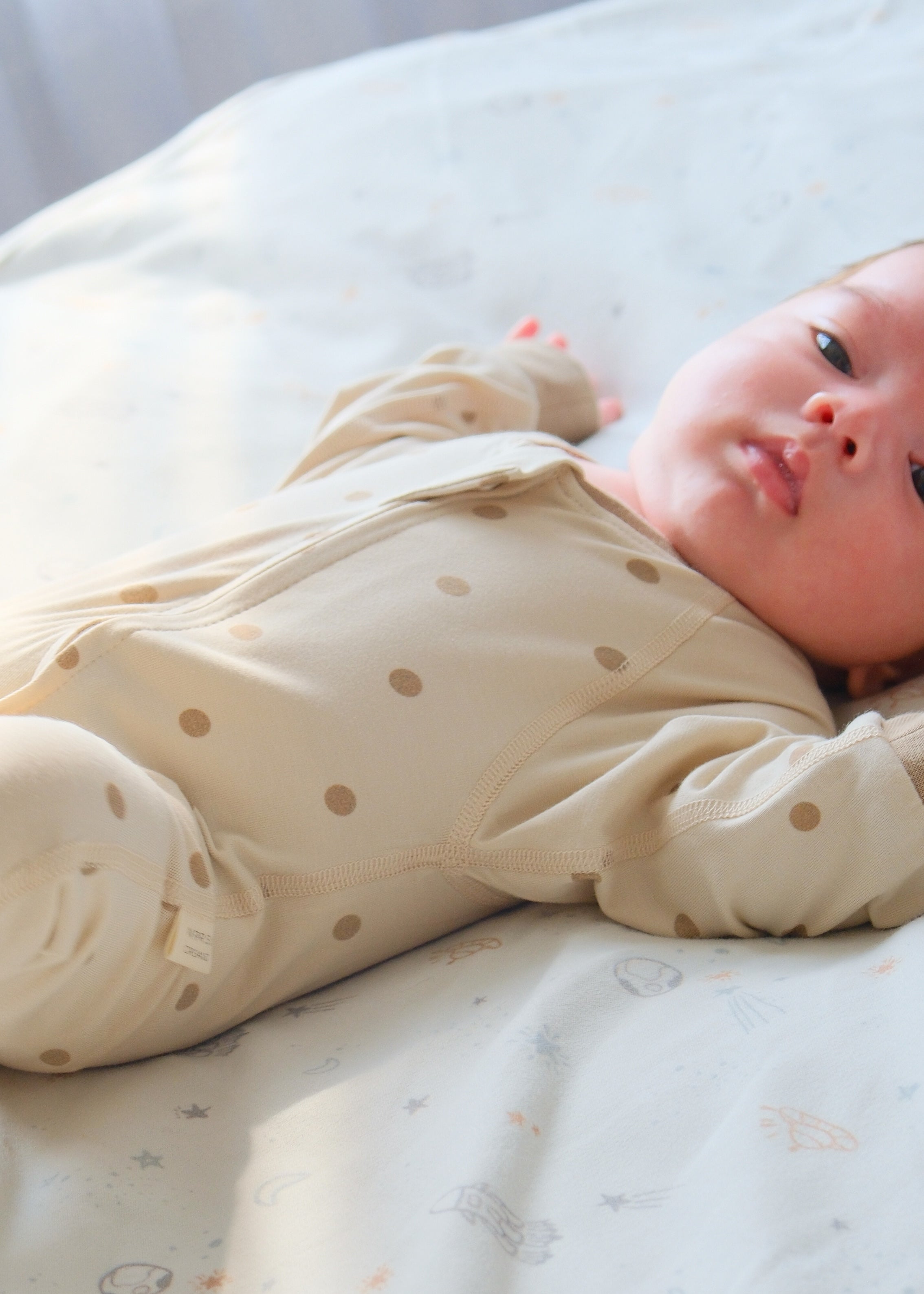 A cute baby peacefully resting on a bed, dressed in an organic bamboo zip-up sleeper with dot patterns