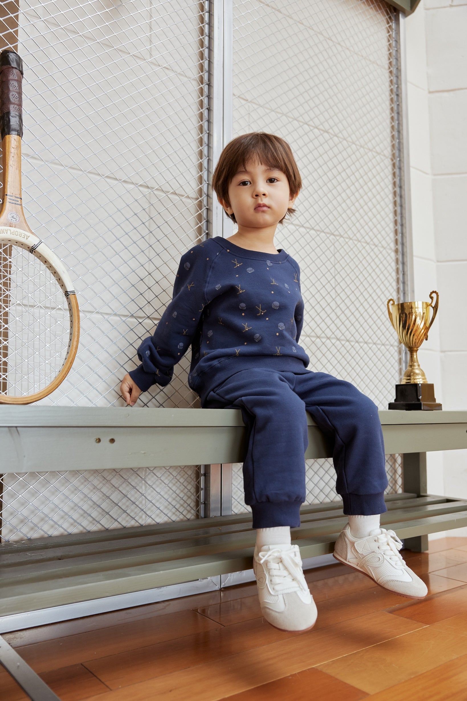 Child sitting on a bench with a tennis racket and trophy in the background