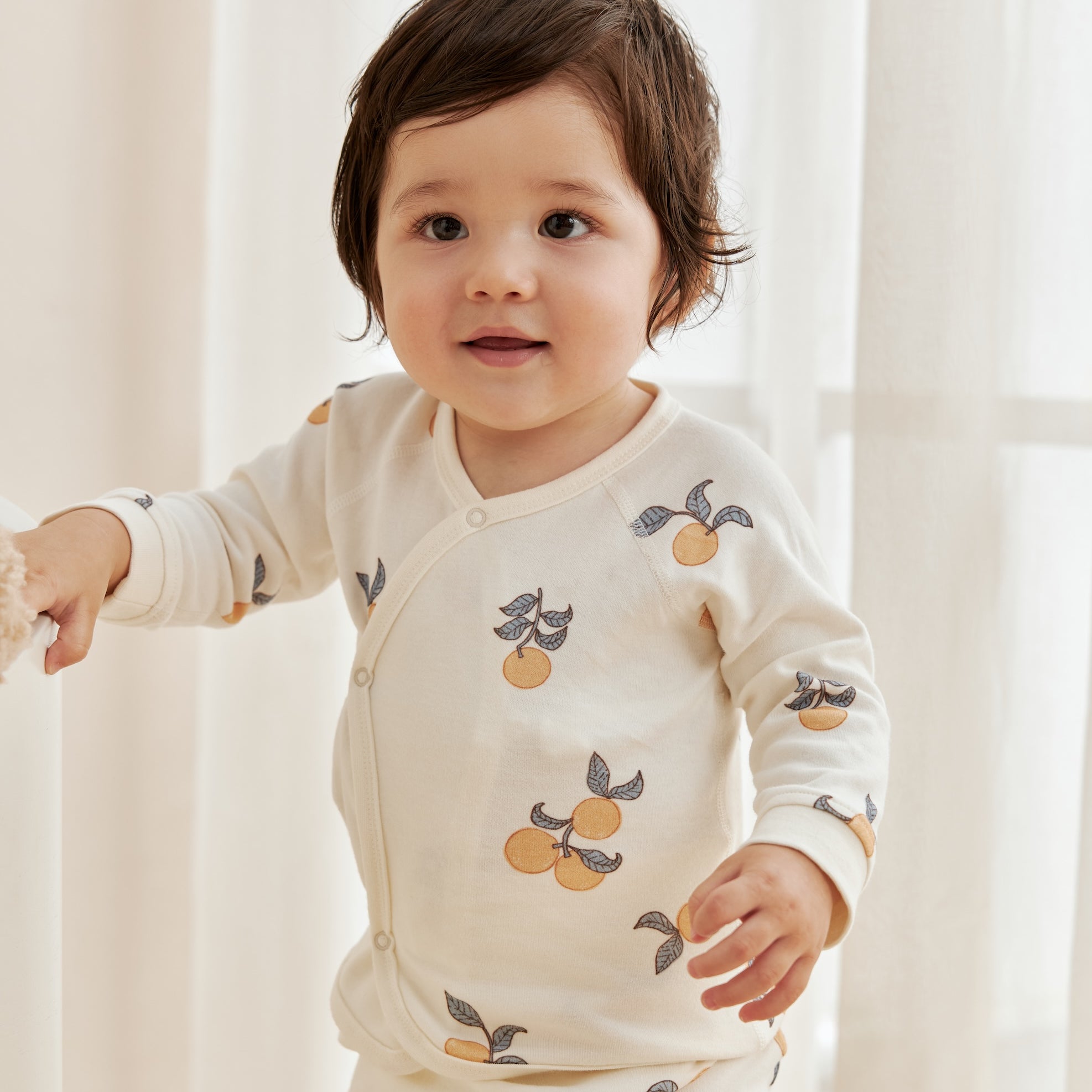 Child wearing a cream-colored outfit with fruit pattern in a softly lit room.