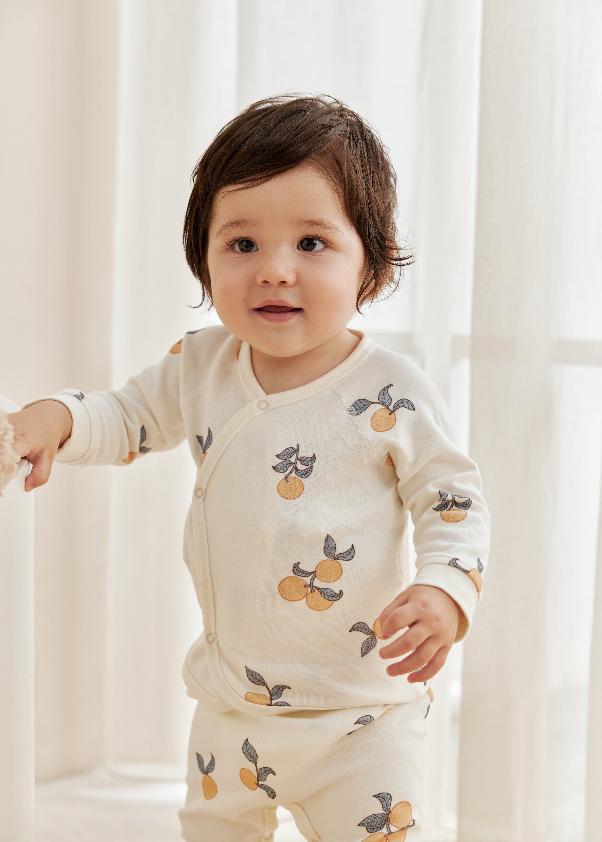 Child wearing a cream-colored outfit with fruit pattern in a softly lit room.