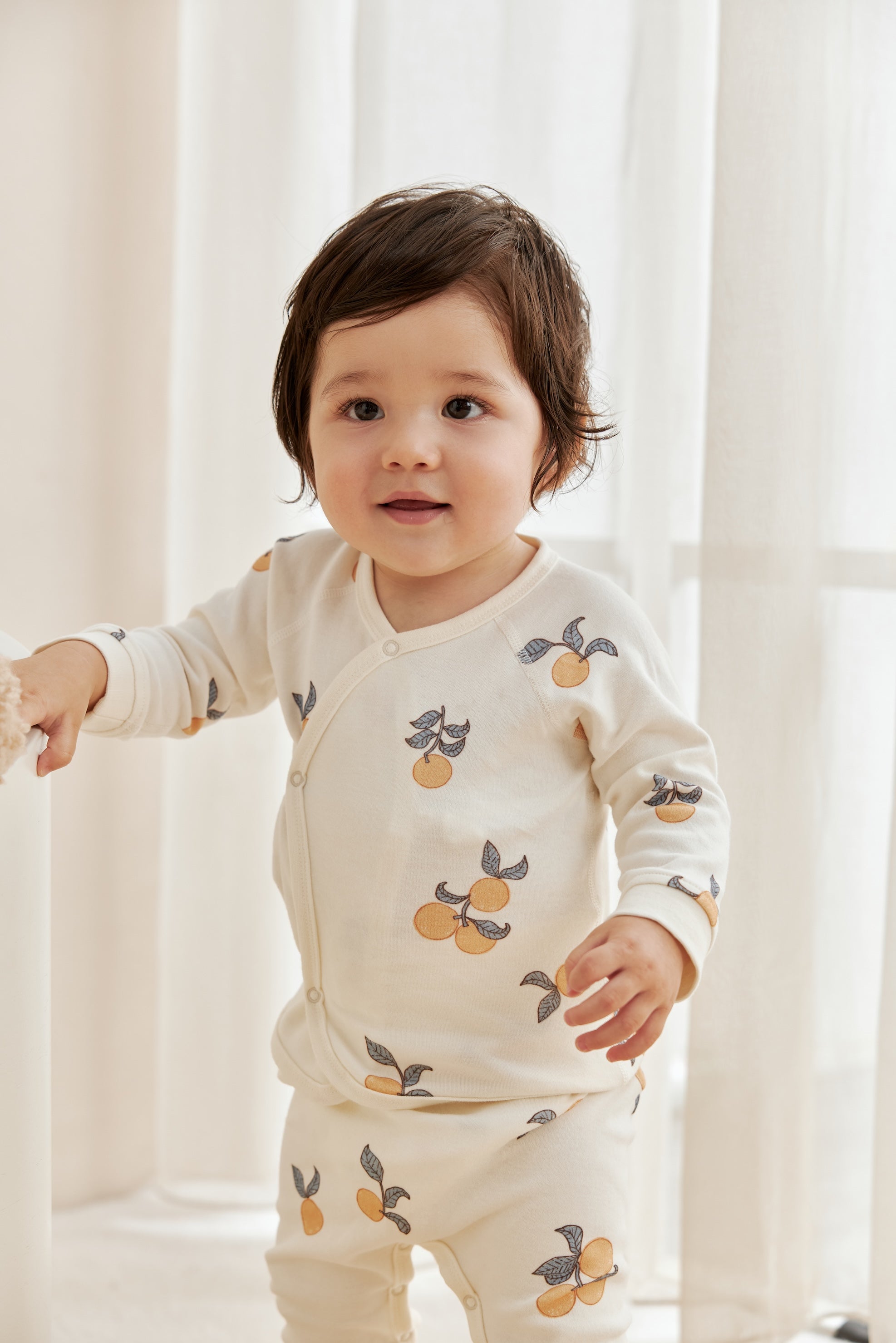 Child wearing a cream-colored outfit with fruit pattern in a softly lit room.