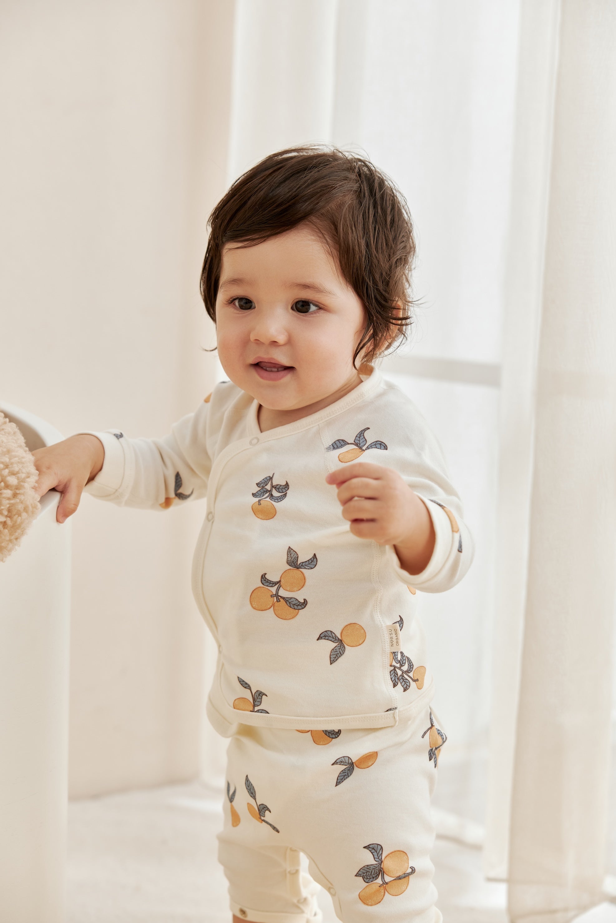 Baby wearing a white outfit with fruit patterns in a softly lit room.