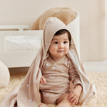 Baby sitting on a carpeted floor wearing a beige hooded cloak.