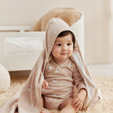 Baby sitting on a carpeted floor wearing a beige hooded cloak.