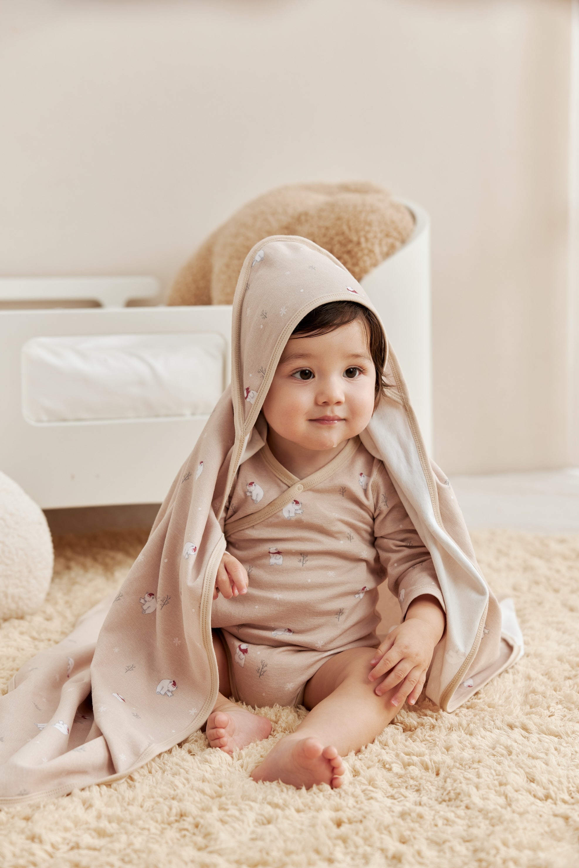 Baby sitting on a carpeted floor wearing a beige hooded cloak.