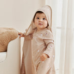 Child wearing a beige hooded outfit in a bright room with white curtains.