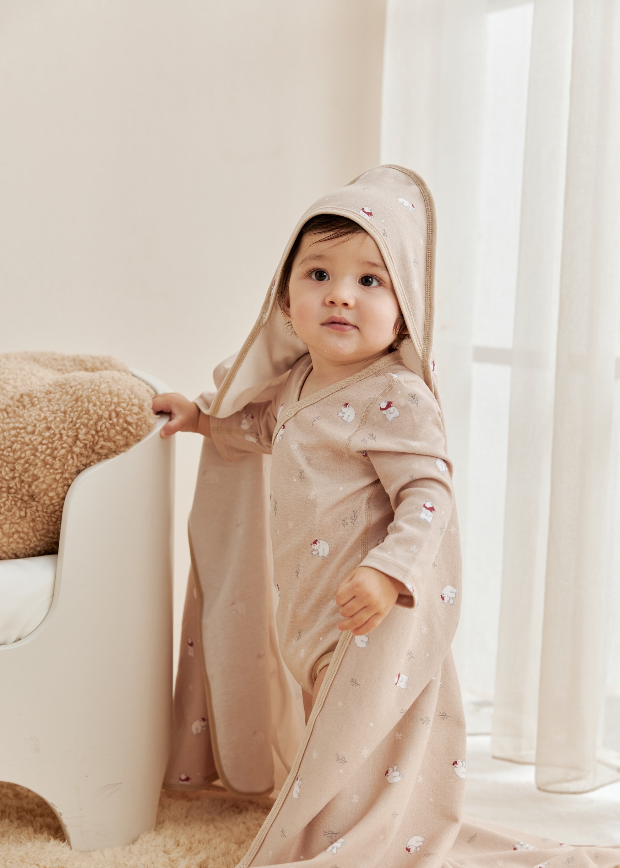 Child wearing a beige hooded outfit in a bright room with white curtains.