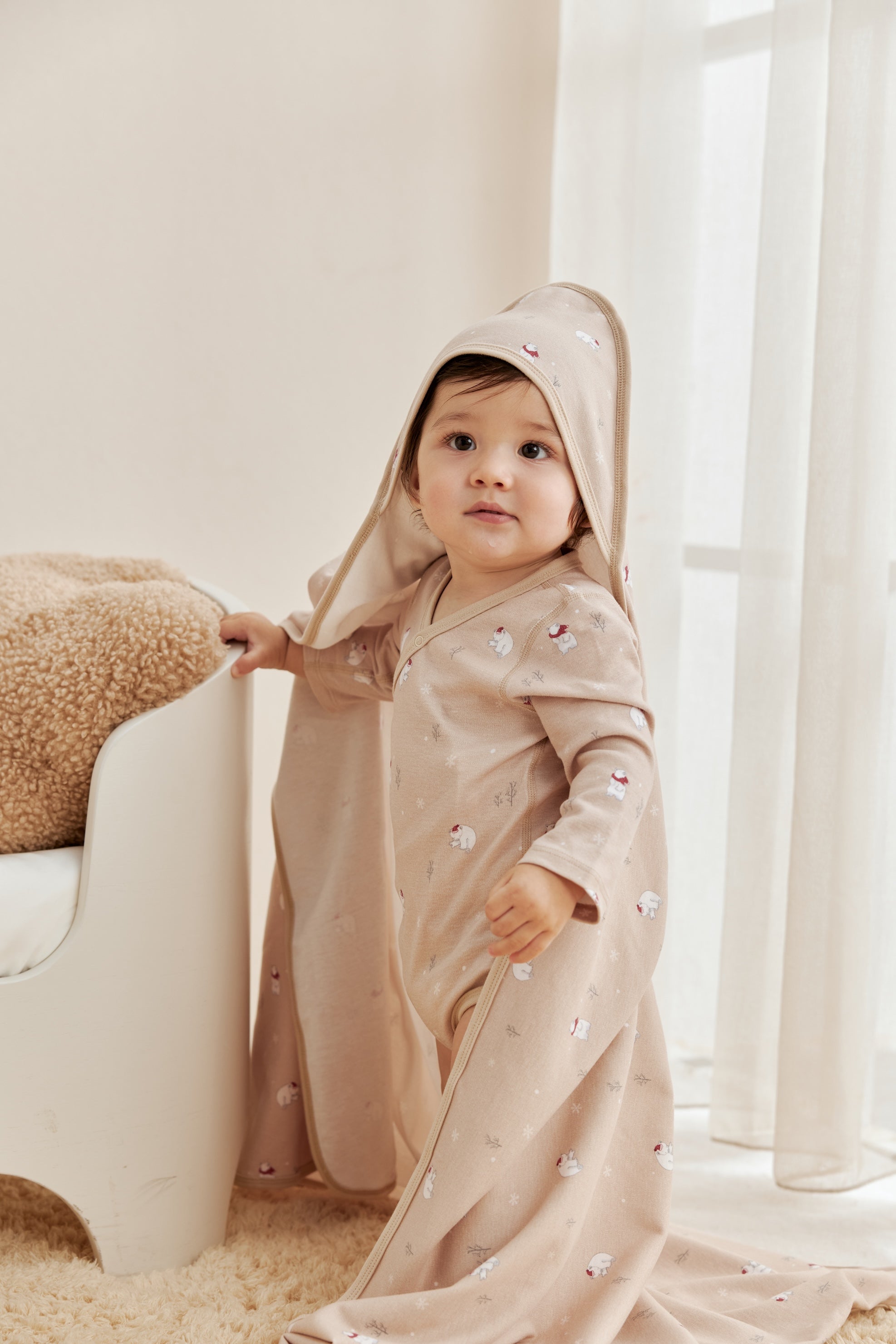 Child wearing a beige hooded outfit in a bright room with white curtains.