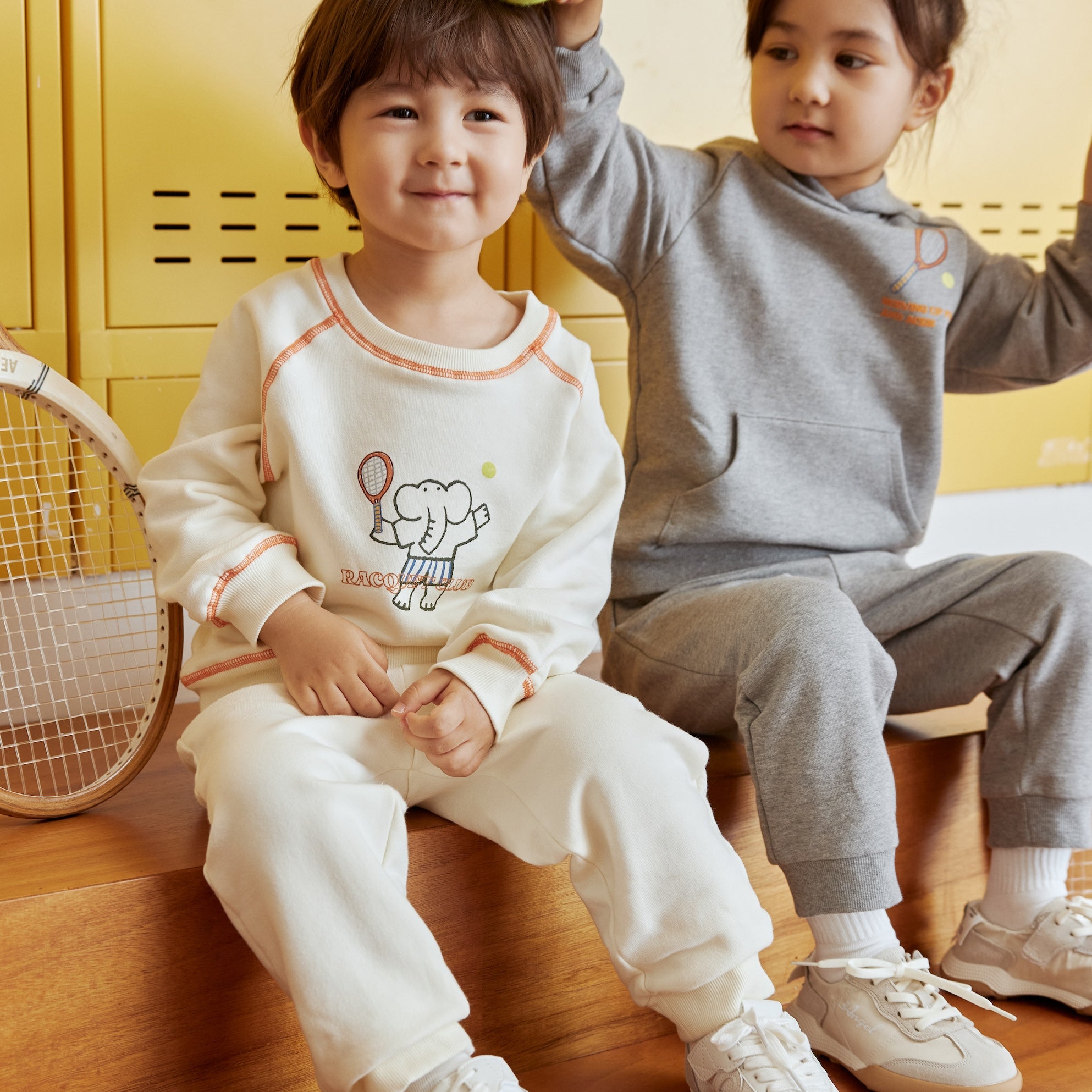 Two children sitting on a wooden floor with a yellow locker in the background.