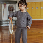 Child in a gray outfit holding a tennis racket in an indoor setting with lockers.