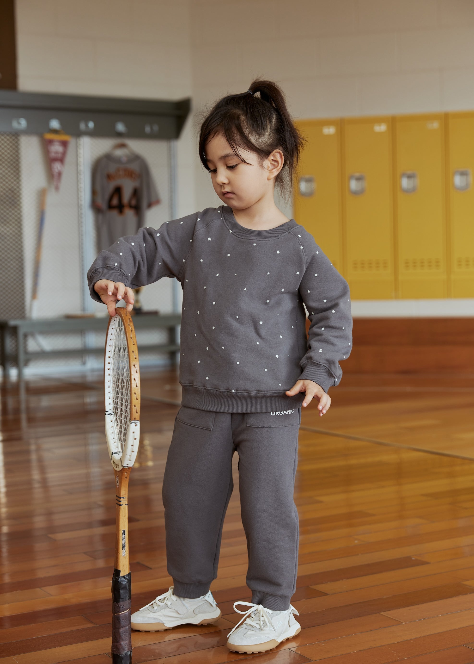 Child in a gray outfit holding a tennis racket in an indoor setting with lockers.