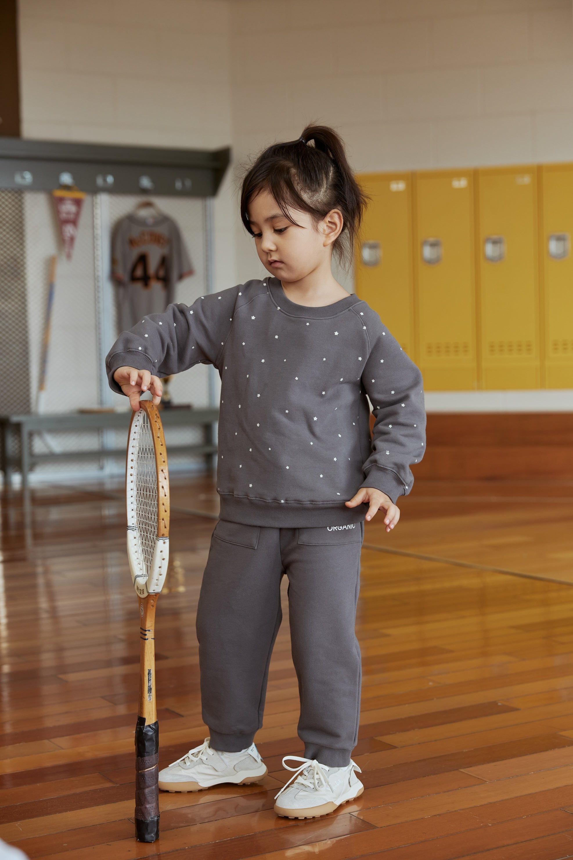 Child in a gray outfit holding a tennis racket in an indoor setting with lockers.