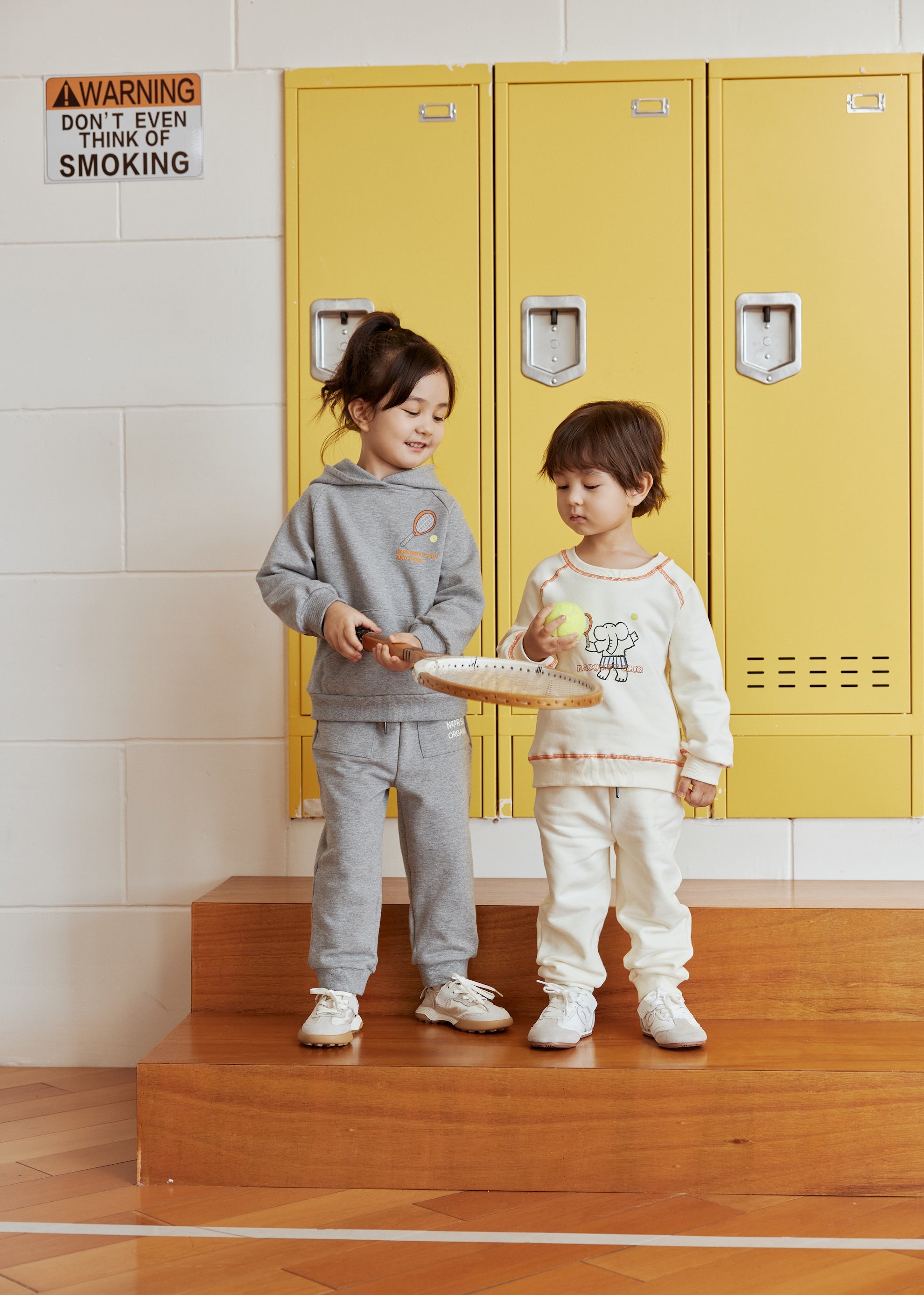 Two children standing on wooden steps in front of yellow lockers with a warning sign above.