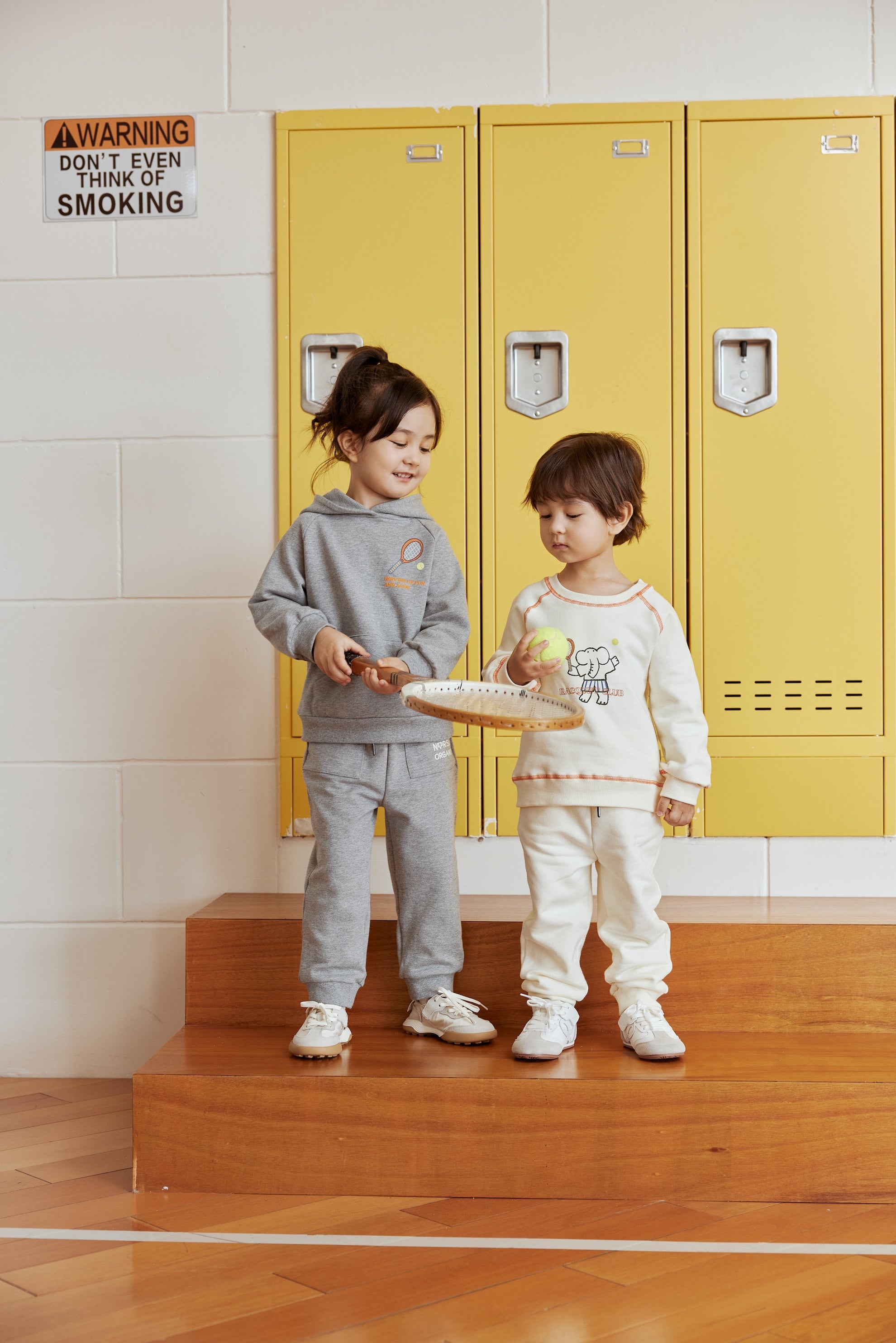 Two children standing on wooden steps in front of yellow lockers with a warning sign above.