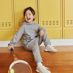 Child sitting on wooden floor with tennis racket and ball, yellow lockers in background