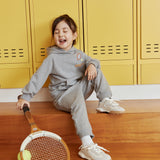 Child sitting on wooden floor with tennis racket and ball, yellow lockers in background