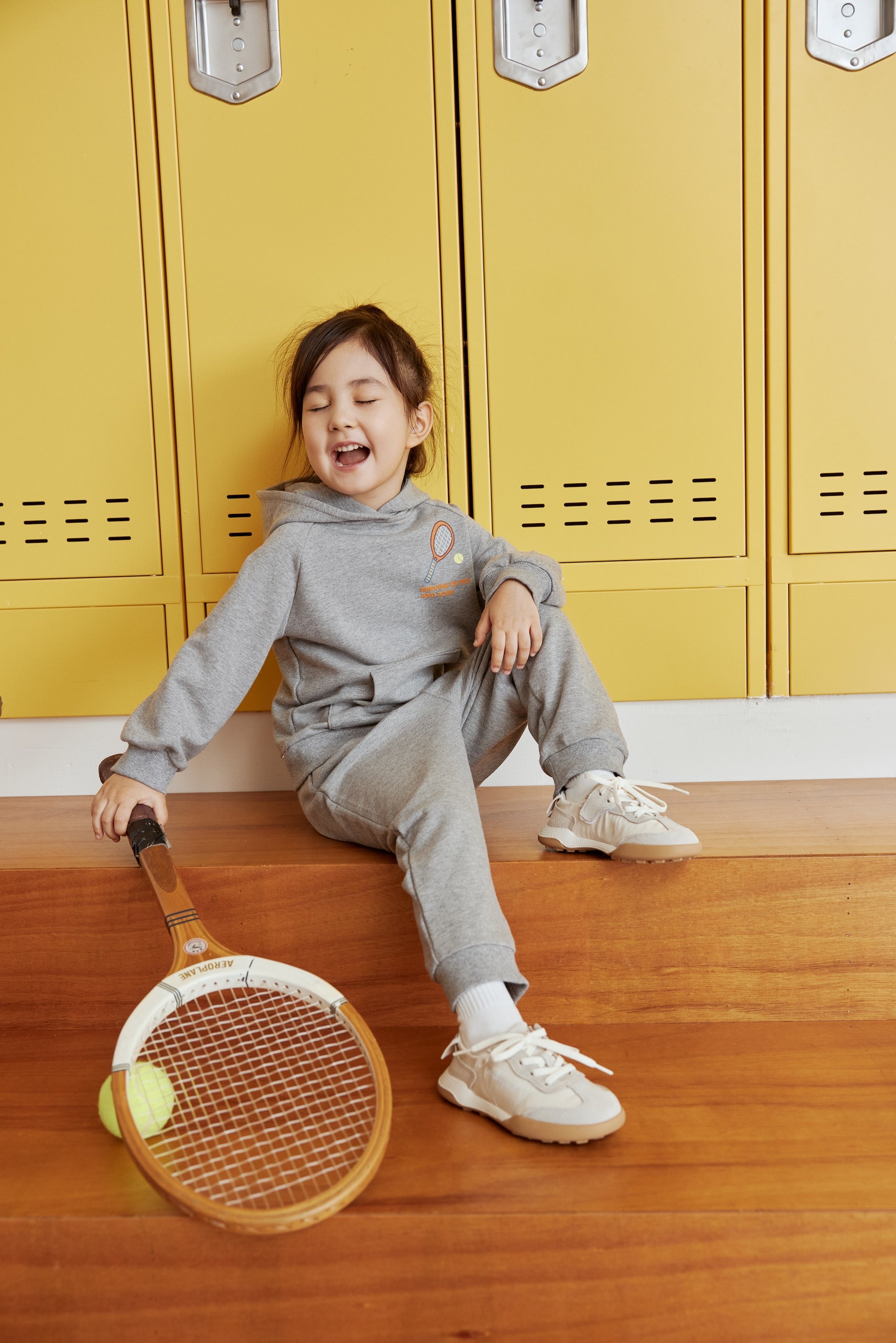 Child sitting on wooden floor with tennis racket and ball, yellow lockers in background