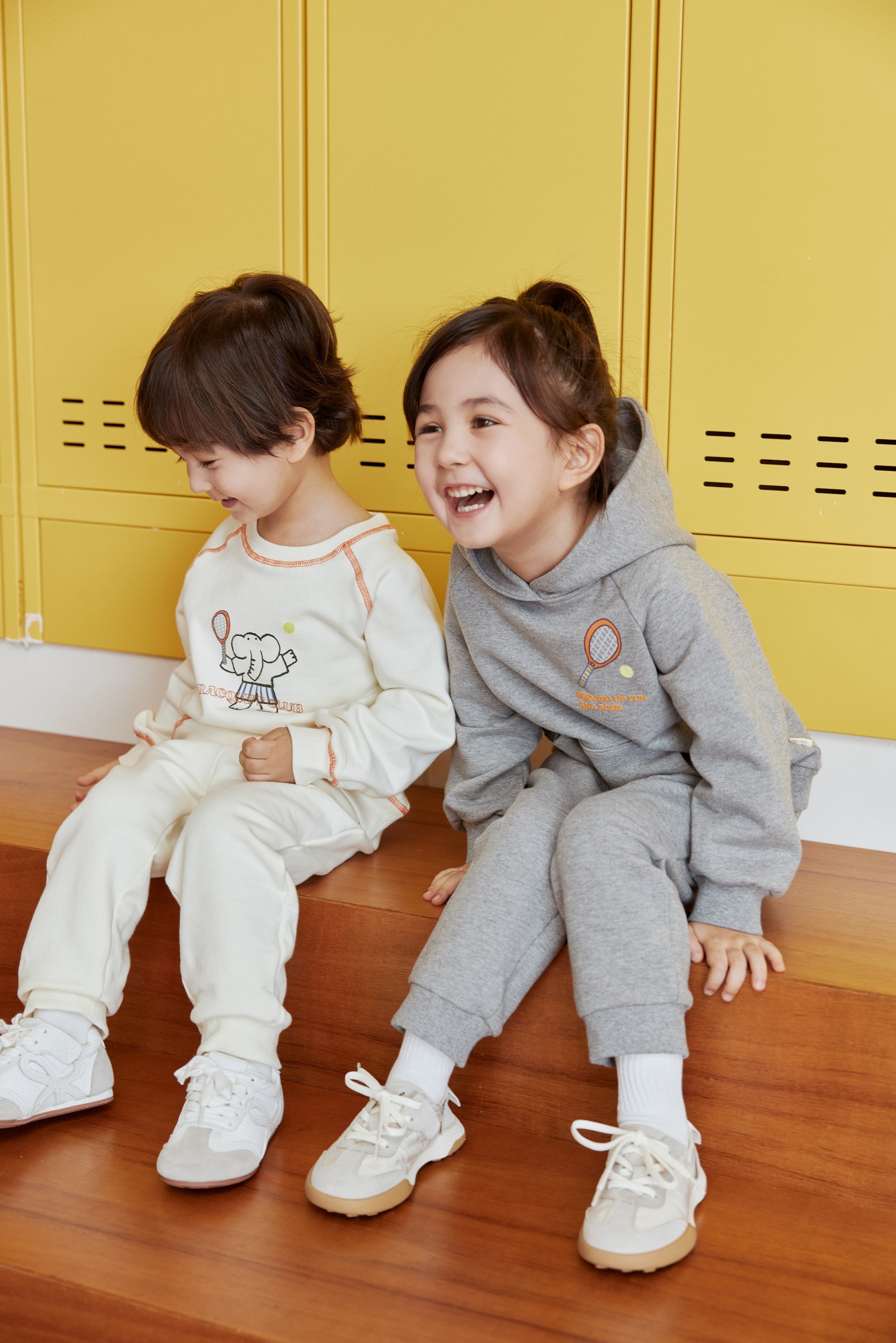Two children sitting on a wooden floor in front of yellow lockers, wearing matching outfits.