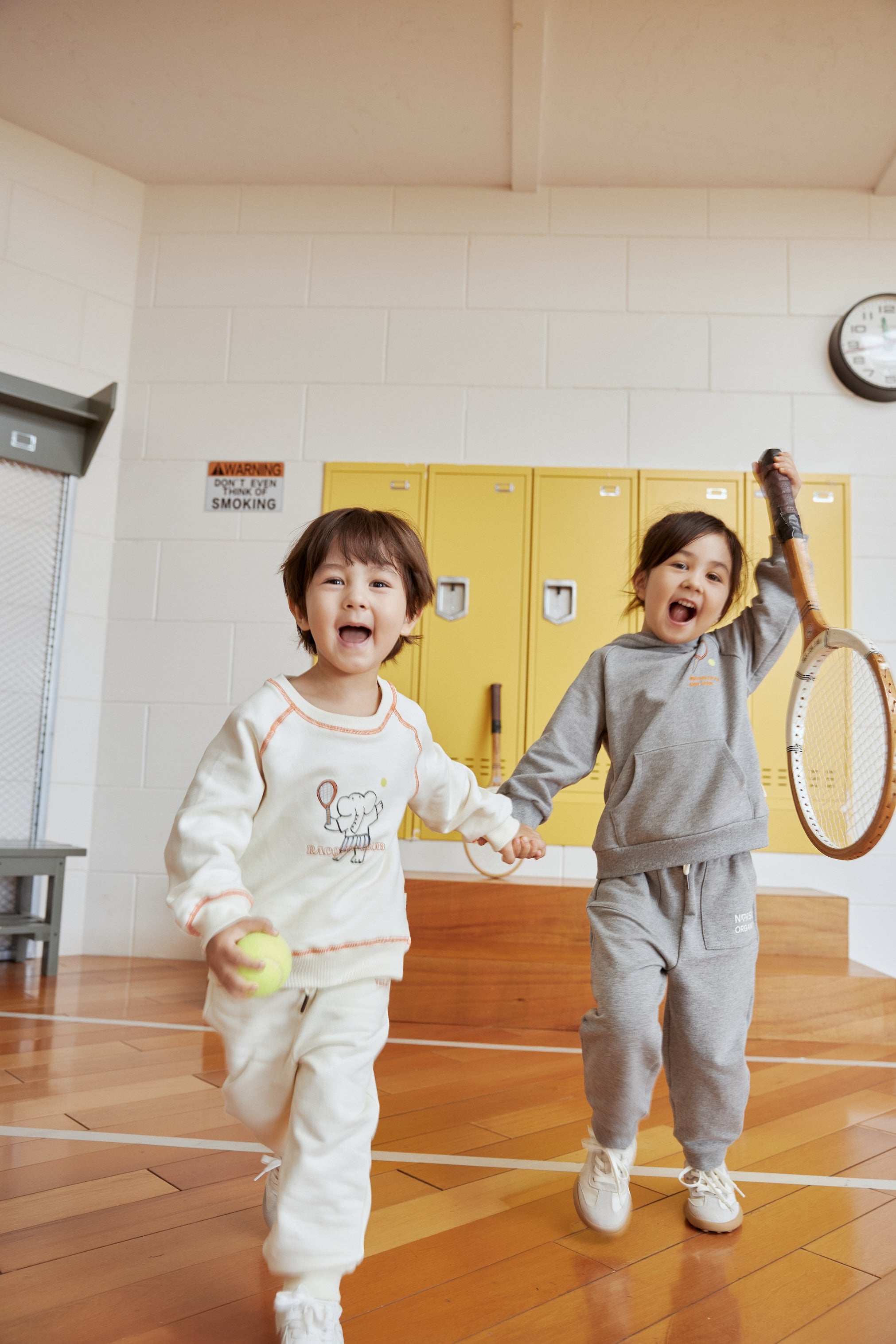 Two children playing with a tennis ball and racket in an indoor setting with yellow lockers.