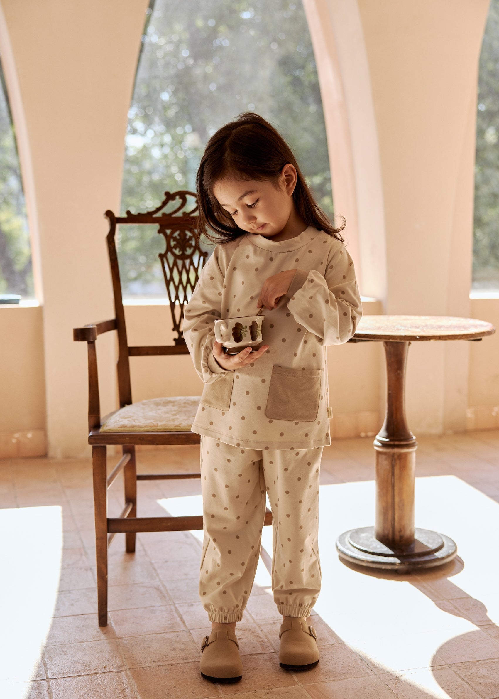 A young girl in a dotted organic bamboo long-sleeve pajama shirt stands playfully in front of a chair