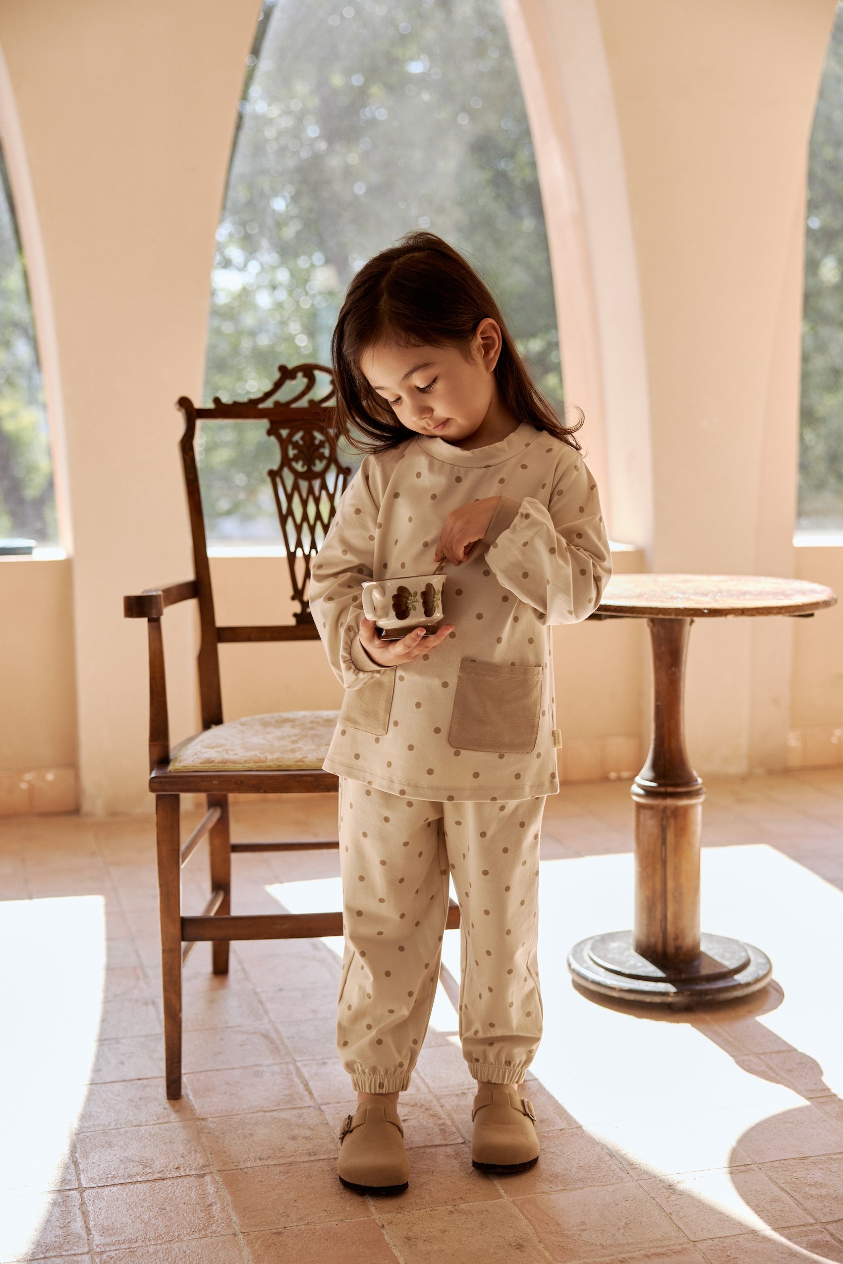 A young girl in a dotted organic bamboo long-sleeve pajama shirt stands playfully in front of a chair