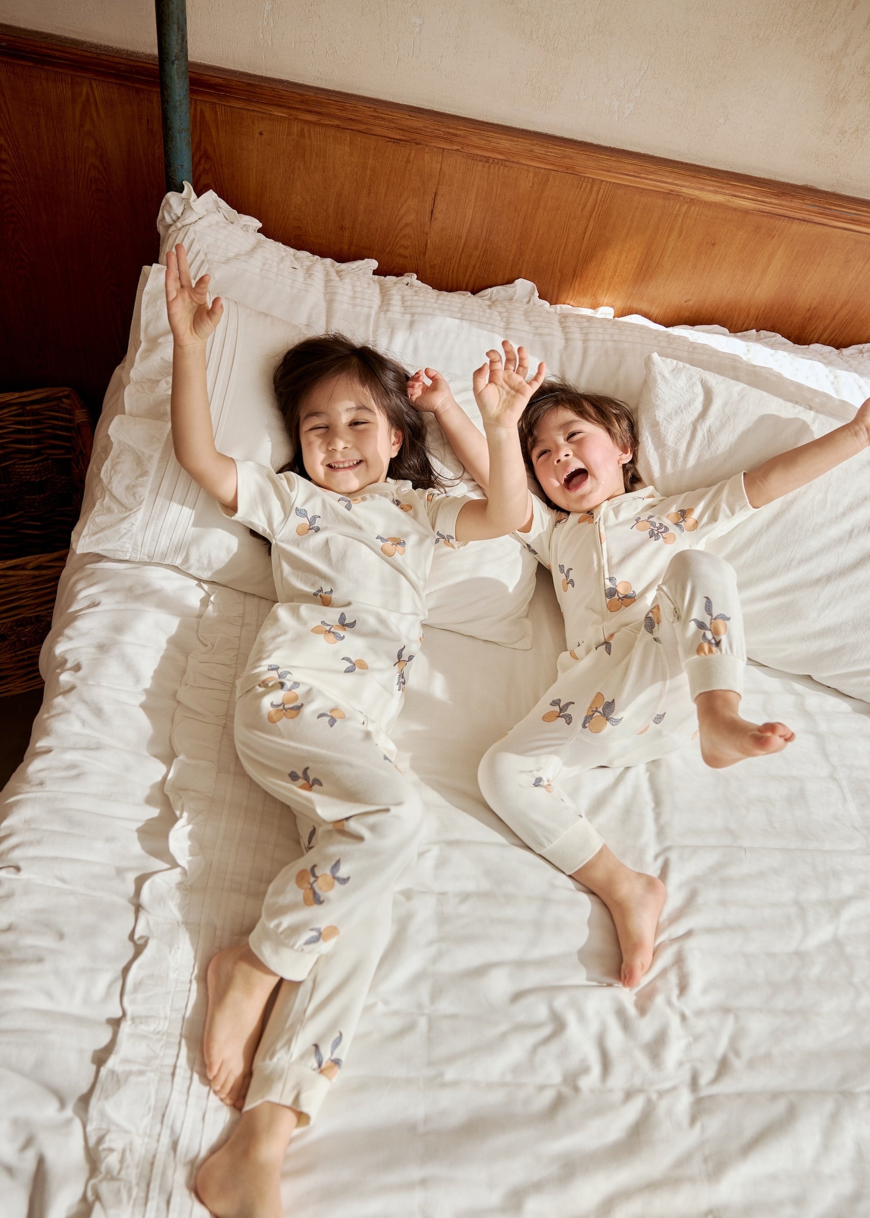 Two kids playing on a bed, one in an organic bamboo pajama set- clementine the other one in an organic zip up sleeper -clementine, embracing the spirit of bedtime fun