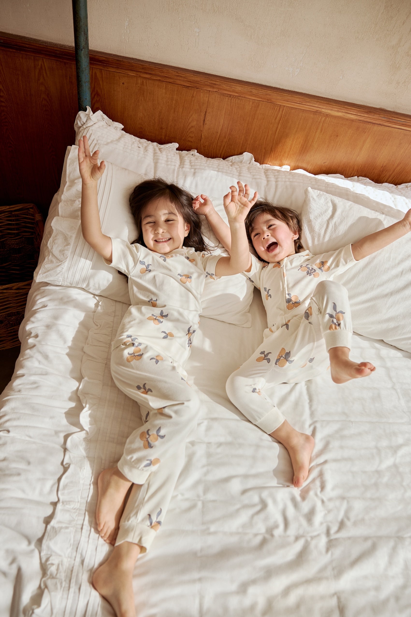 Two kids playing on a bed, one in an organic bamboo pajama set- clementine the other one in an organic zip up sleeper -clementine, embracing the spirit of bedtime fun