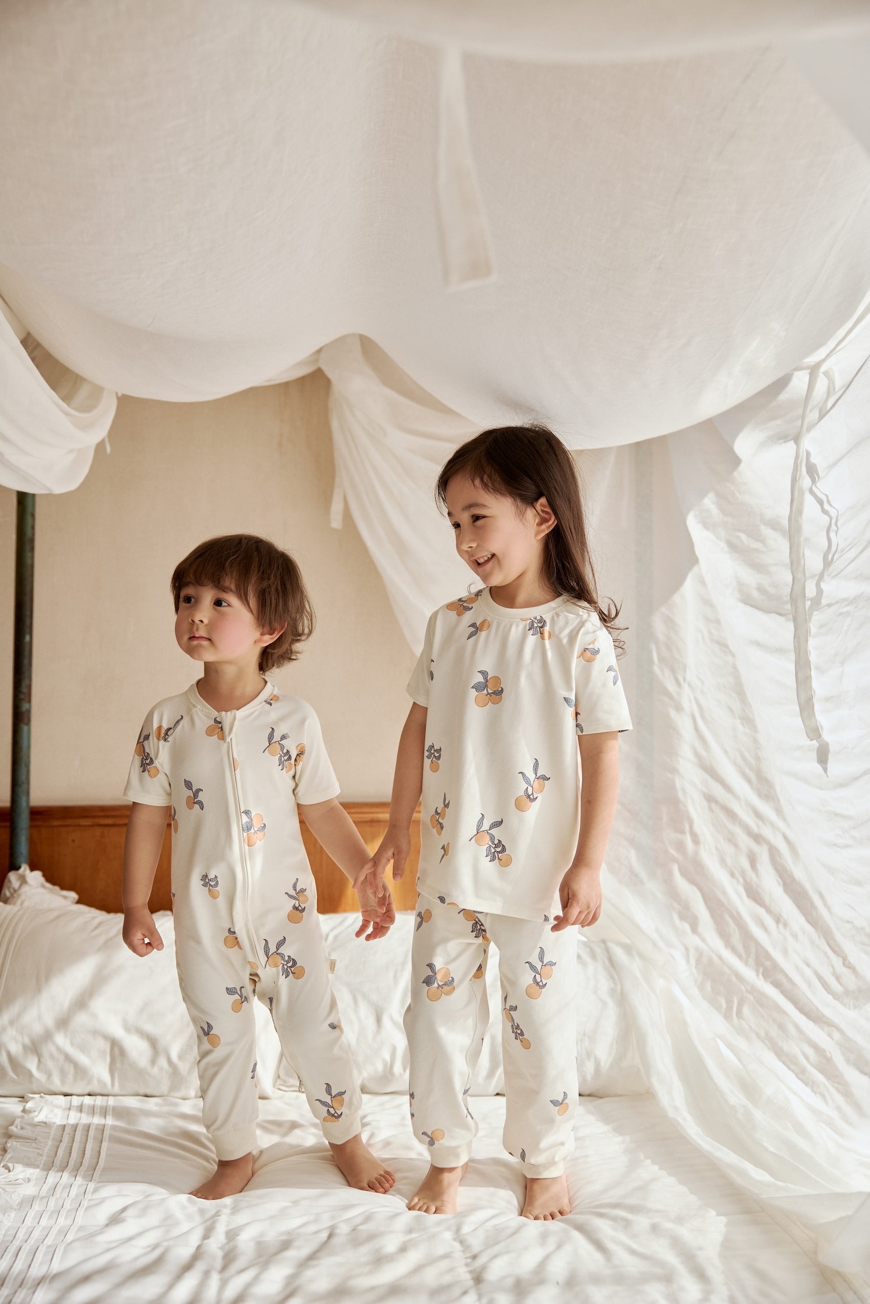 Two kids standing on a bed, one in an organic bamboo pajama set- clementine the other one in an organic zip up sleeper -clementine, embracing the spirit of bedtime fun