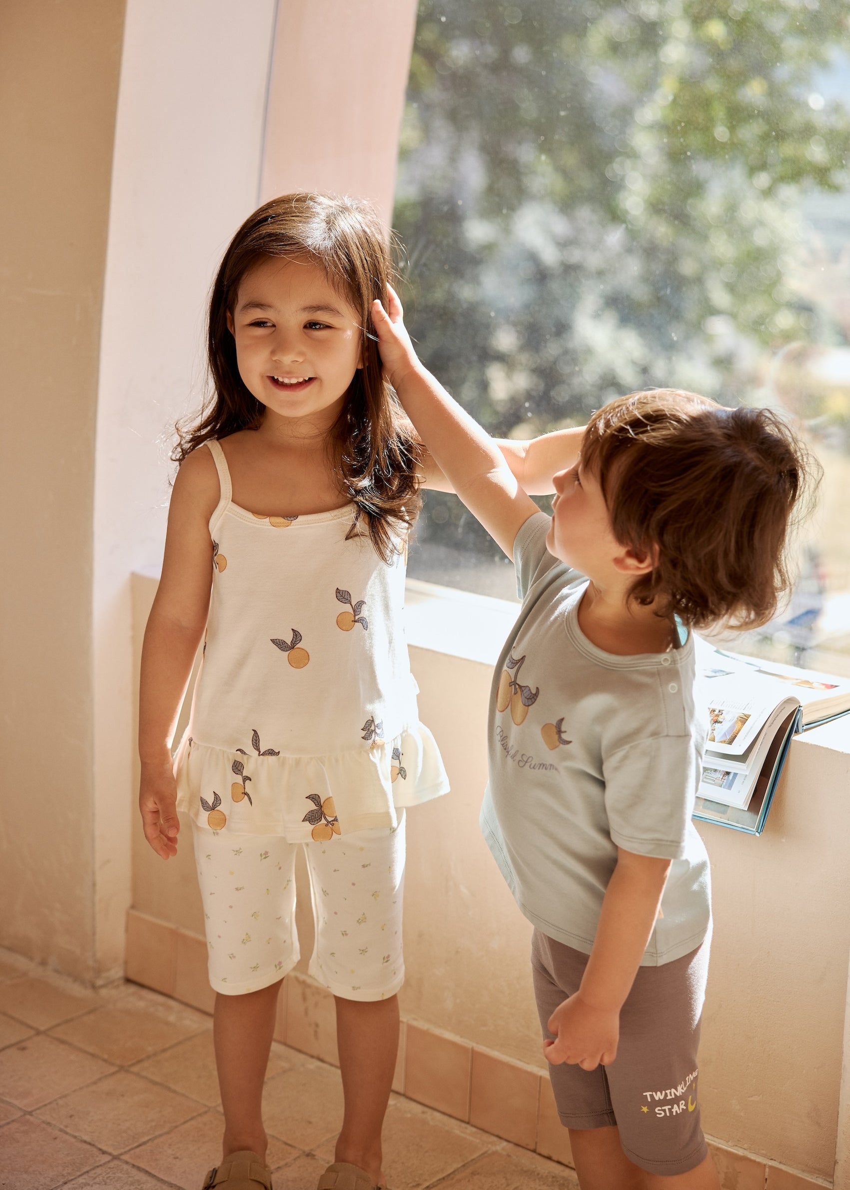 Two children standing by a sunny window, smiling and interacting, wearing light-colored outfits with fruit prints
