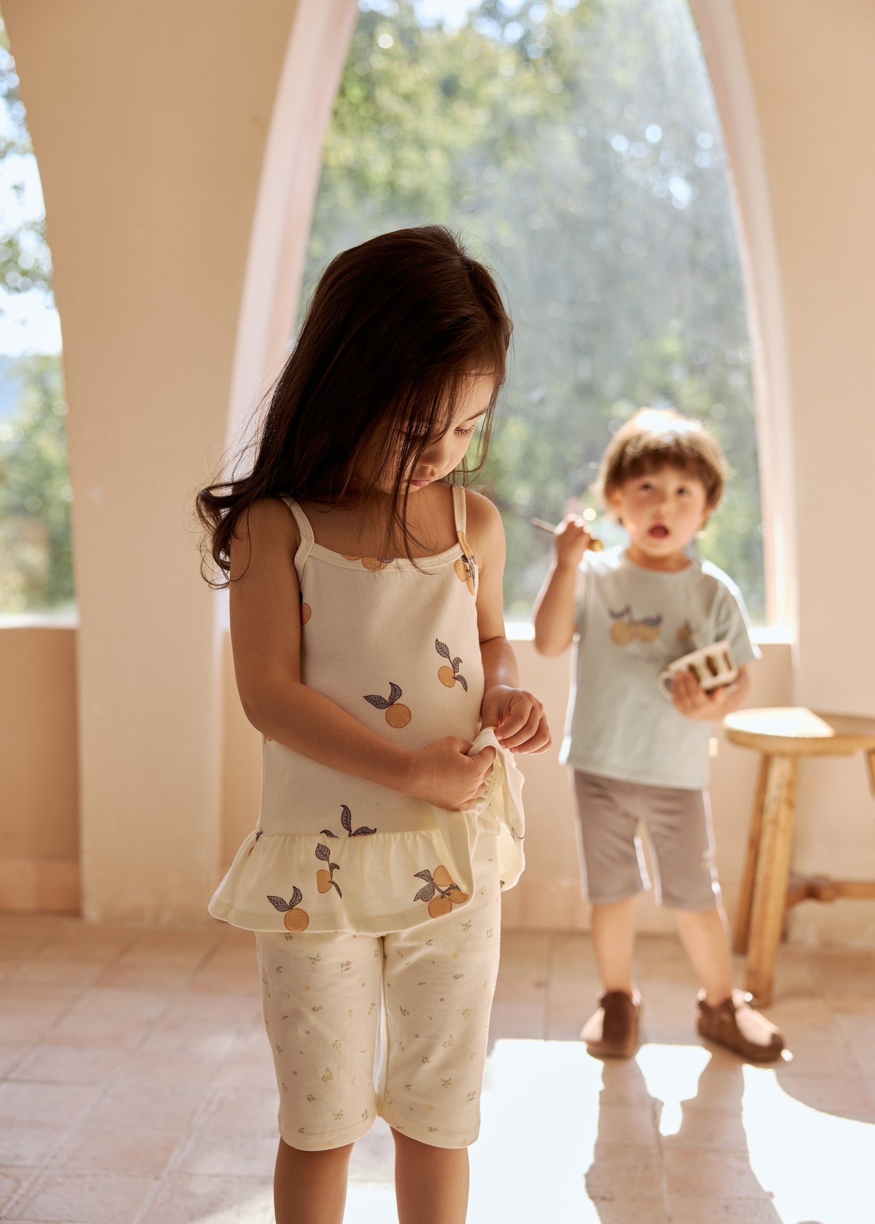Two children in a sunlit room, the girl in the foreground looks down at her outfit, while the boy in the background holds a small toy and looks forward