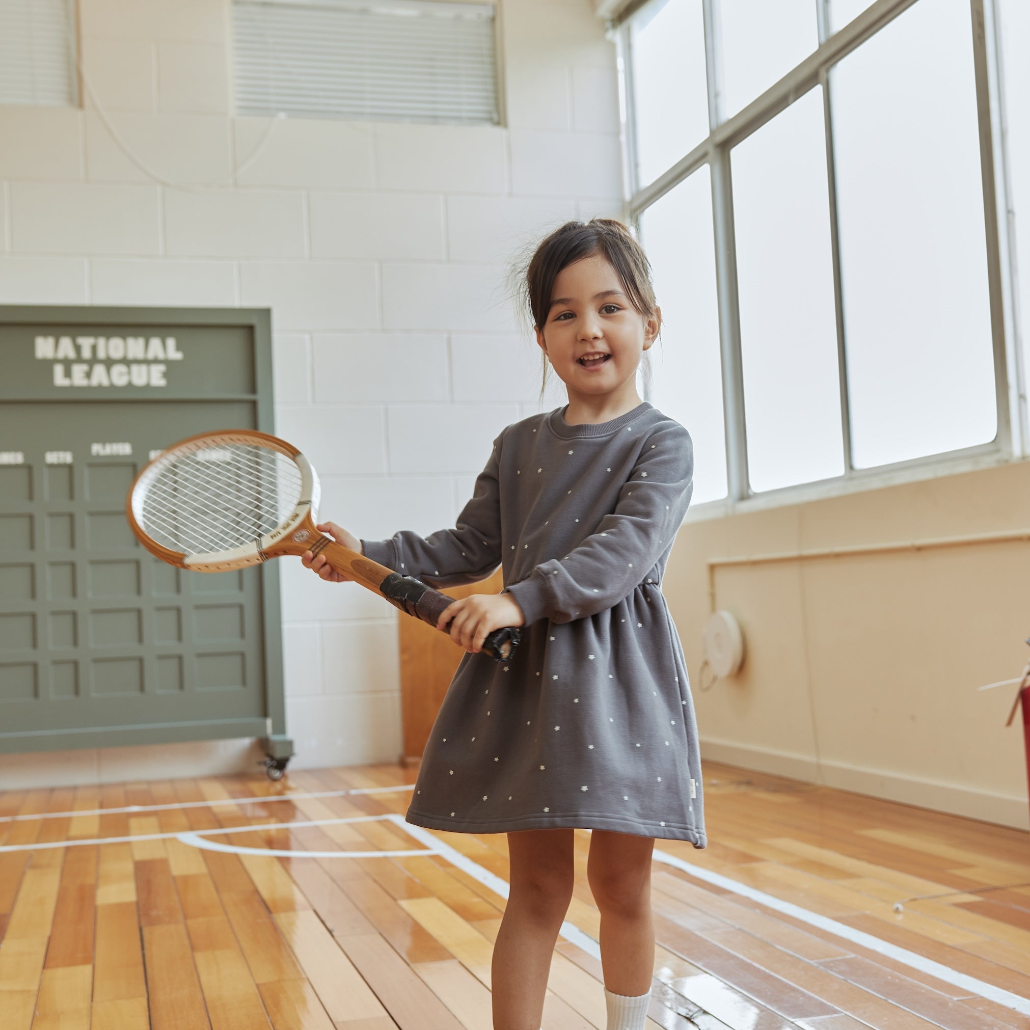 Young girl in a gray dress holding a tennis racket indoors.