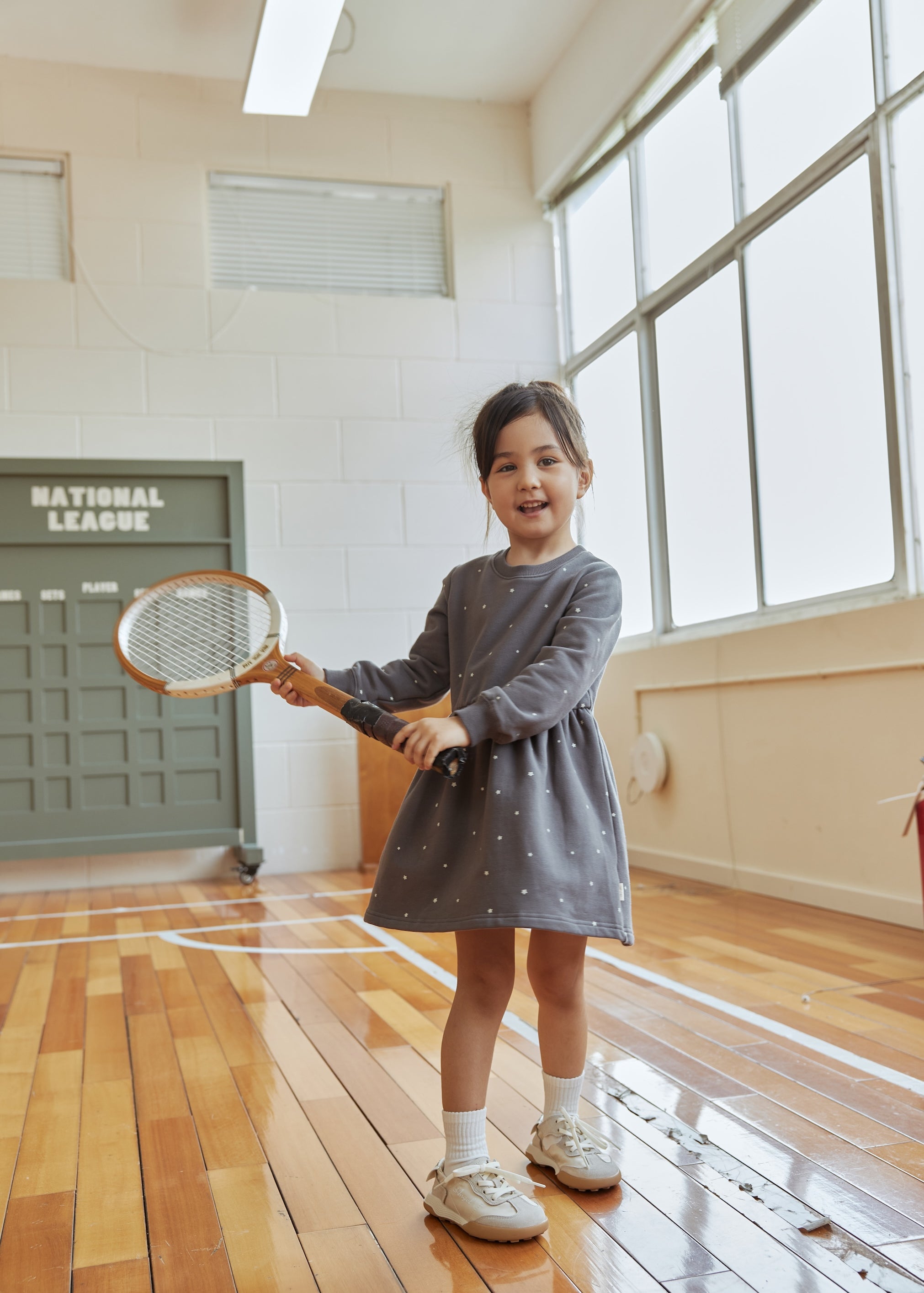 Young girl in a gray dress holding a tennis racket indoors.